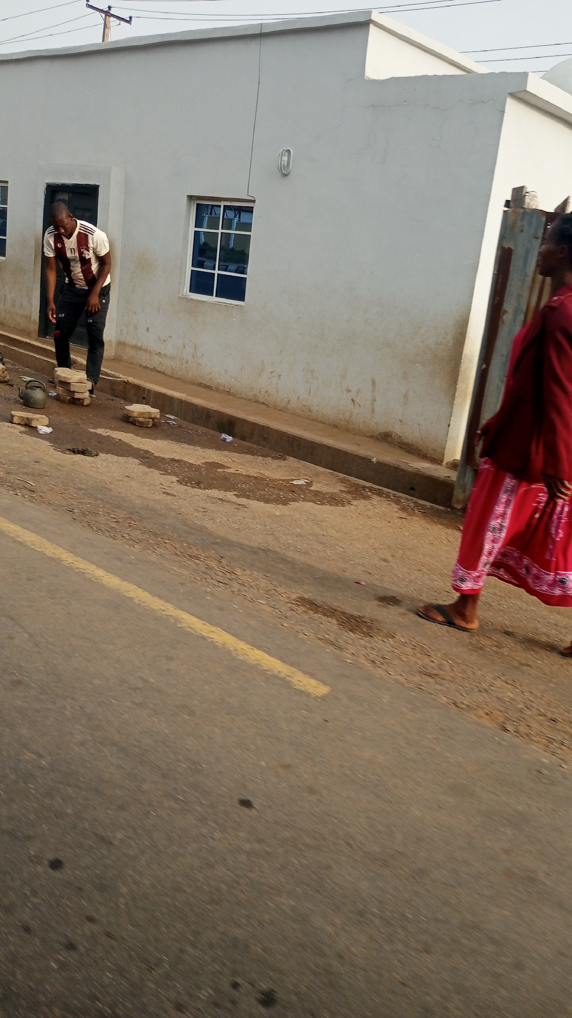 The image depicts a daytime street scene in Adankolo, Nigeria, featuring a man, a partially visible woman, and a light-colored building.
On the left side of the frame, a man is captured in mid-action, seemingly stepping down from a raised area or curb. He wears a white short-sleeved shirt with maroon and white horizontal stripes and dark trousers. His gaze is directed downwards. Nearby, on the dirt ground next to the curb, are several rectangular, light-colored paver blocks and a dark, squat kettle or pot.
To the right, partially cut off by the frame, a woman walks away from the viewer. She wears a long-sleeved, deep red top and a flowing, long red skirt adorned with white patterns along the hem. She has black sandals on her feet.
Behind the man and extending across the background is a simple, single-story building with light grey or off-white plastered walls. It has a dark doorway and two prominent windows with bright blue panes or frames, reflecting the sky. A white, oval-shaped light fixture is mounted on the wall between the door and the rightmost window. Utility poles and power lines are visible against the sky above the building.
The foreground is dominated by a grey asphalt road with a single, faded yellow line running down its length. A concrete curb separates the road from a rough, dirt-strewn area where the man stands, containing some litter. The lighting suggests it is daytime, possibly morning or late afternoon, with soft, diffused light and subtle shadows. No discernible text is visible on any objects or individuals. The overall impression is an everyday street view in a residential or semi-urban setting.