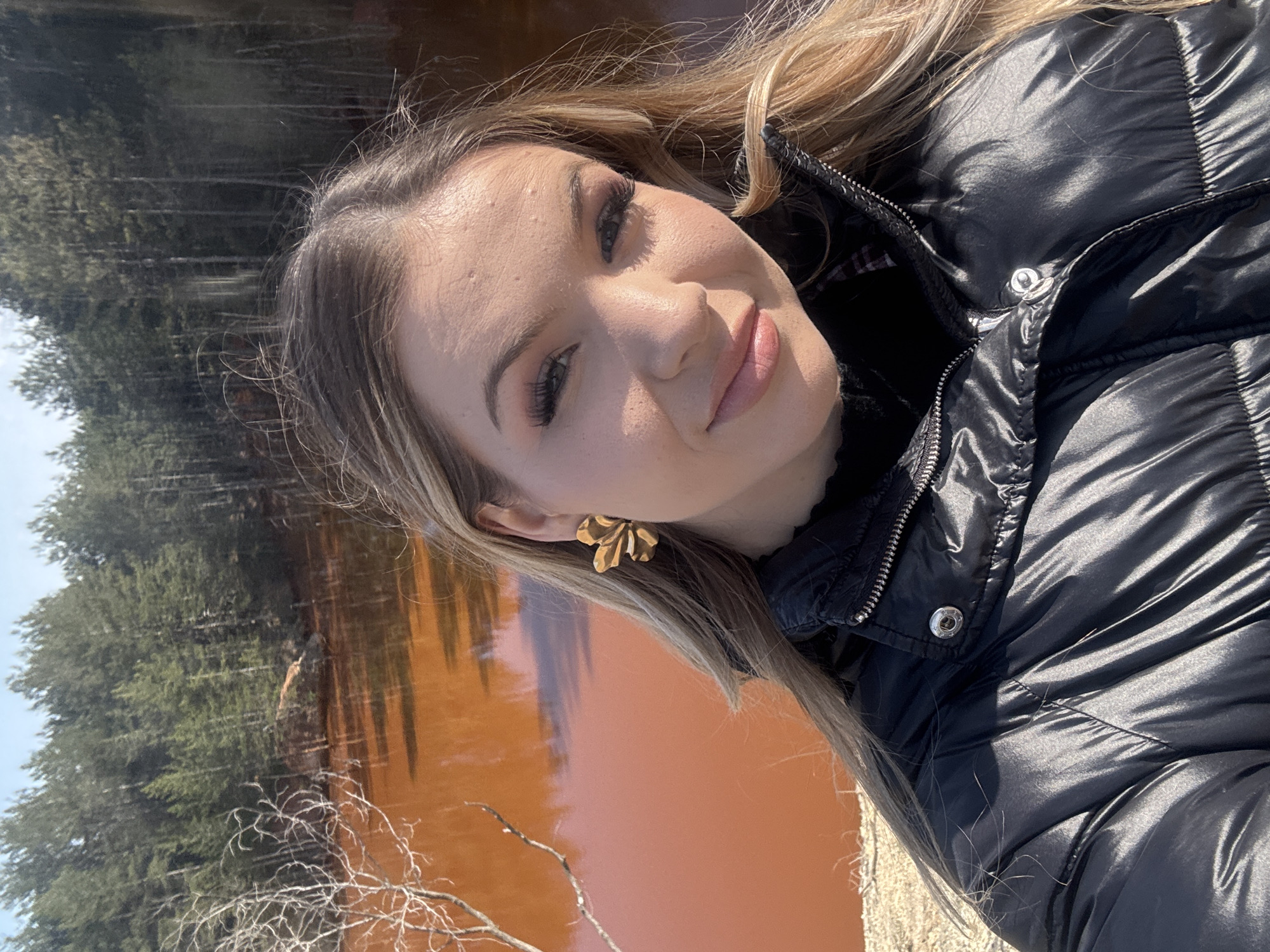 A woman takes a selfie outdoors. The background consists of a rust-colored body of water bordered by coniferous trees. The water and trees suggest the scene is located near the abandoned Elshitsa copper mine in Elshitsa, Bulgaria, where the water is polluted by iron oxides and heavy metals. The woman is wearing a black puffer jacket and gold floral earrings. The light indicates it is daytime.