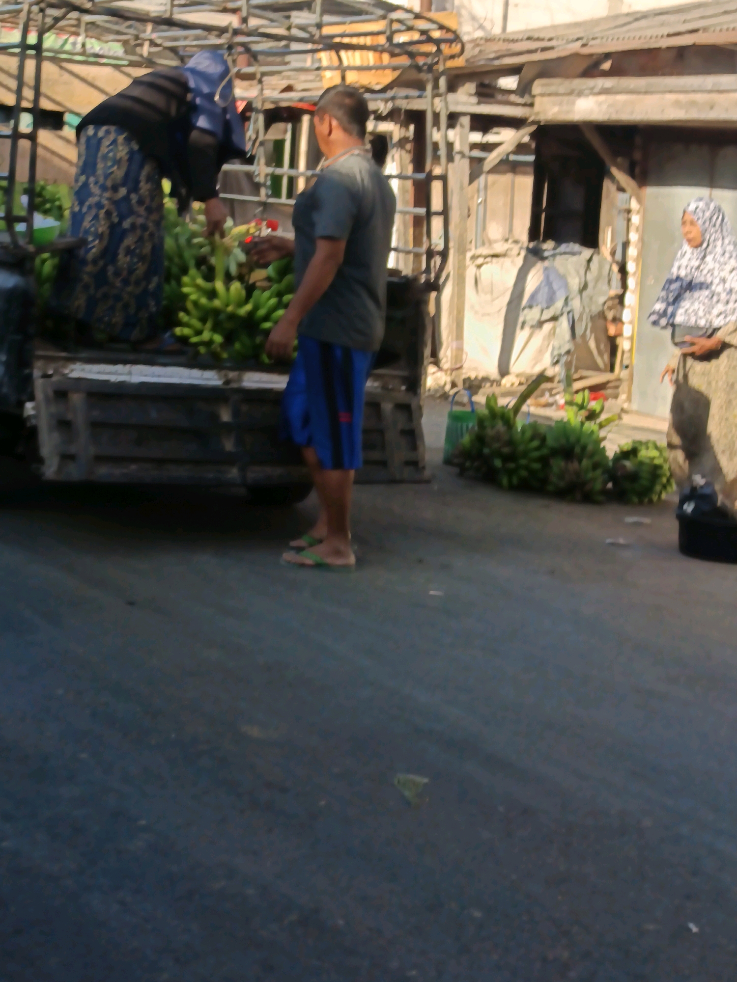 In the city of Blega, Indonesia, on a paved street, two women and one man are present near a flatbed truck. The woman on the left, wearing a dark hijab and a blue and gold patterned skirt, is leaning over the truck, potentially arranging or displaying bananas. Next to her, a man in a grey t-shirt and blue shorts stands facing the woman, his body angled towards the truck's contents. To the right of the scene, a second woman, wearing a patterned hijab and a light-colored garment, stands holding a black container. Several bunches of bananas are visible on the ground near the truck, some in a green crate. The background features a rustic building with a corrugated metal roof and wooden structures. The scene appears to be a street market or a point of sale for agricultural produce.