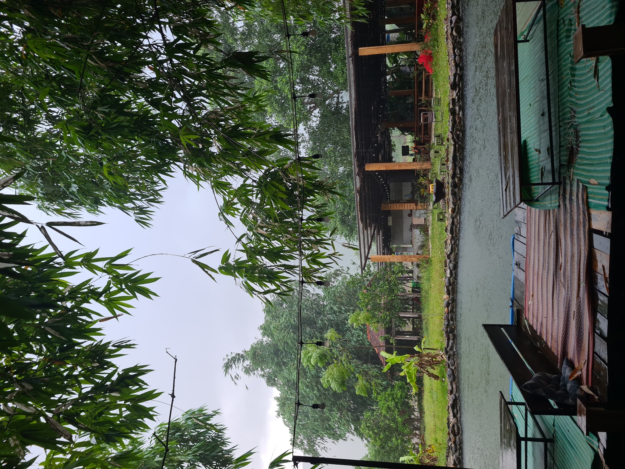 The image depicts an outdoor seating area by a body of water during a rain shower. In the foreground, wooden platforms are visible with tables and seating. One platform has a green mat and a wooden table, while another features a striped mat and a dark wooden table. The water appears choppy due to the rain. Beyond the water, a grassy area is bordered by rocks. A covered structure with wooden pillars and a dark roof is situated behind the grassy area, furnished with tables and chairs. Trees, including bamboo, are abundant, with some branches hanging over the scene and string lights with bare bulbs strung across. A small red-flowered plant is visible near the covered structure. The sky is overcast, indicating a rainy day. The location is implied to be a restaurant or cafe in the city of Đà Nẵng, Vietnam, given the naturalistic outdoor setting commonly found in such establishments in the region. There are no people visible in the image.