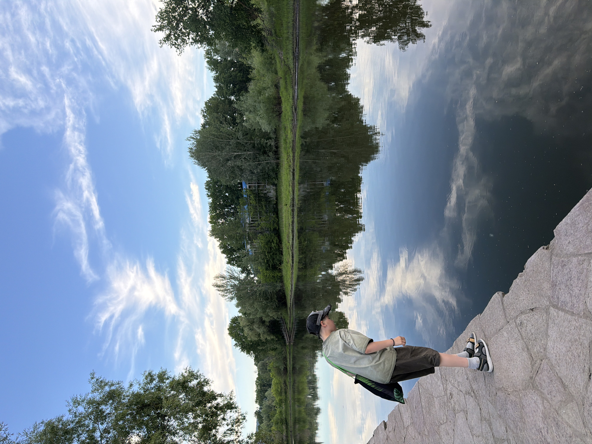 The image shows a young boy wearing a green t-shirt, dark shorts, a baseball cap, and sandals, standing on a stone embankment next to a calm body of water. The water reflects the sky, trees, and clouds, creating a mirror-like effect. The boy is looking out at the water, and his posture suggests he is observing the scene.
The setting appears to be a park or a natural area with lush green trees lining the opposite bank of the water. The sky is a bright blue with scattered white, wispy clouds, indicating pleasant weather. The time of day is likely late afternoon or early evening, given the soft lighting. The image is oriented vertically, with the boy and the embankment at the bottom, and the sky and reflection dominating the upper portion.
Notable details include the texture of the stone embankment and the ripples on the water’s surface. The boy has a bag slung over his shoulder. The overall mood is peaceful and contemplative, with the boy immersed in the serene environment. There is no visible text in the image. The context provided indicates the location is Volzhskiy, Russia.