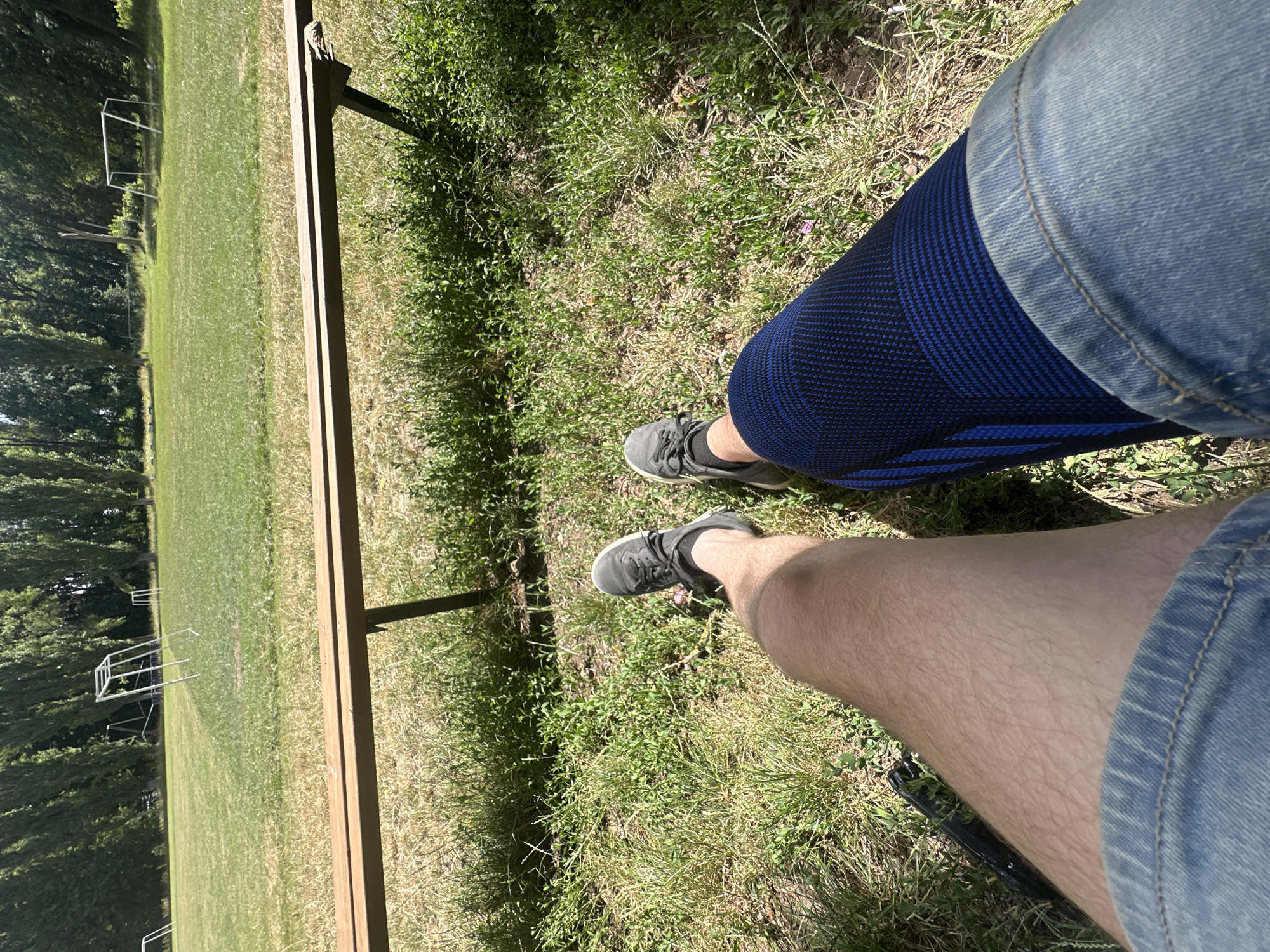 In Makiv, Ukraine, on a sunny day, a person sits on the grass by a soccer field. The person is wearing denim shorts, a blue and black knee brace on their right leg, and grey sneakers. Their left leg is bare and shows hair. The field is green with some brown patches, and two soccer goals are visible in the background. A wooden bench is in front of the person.
