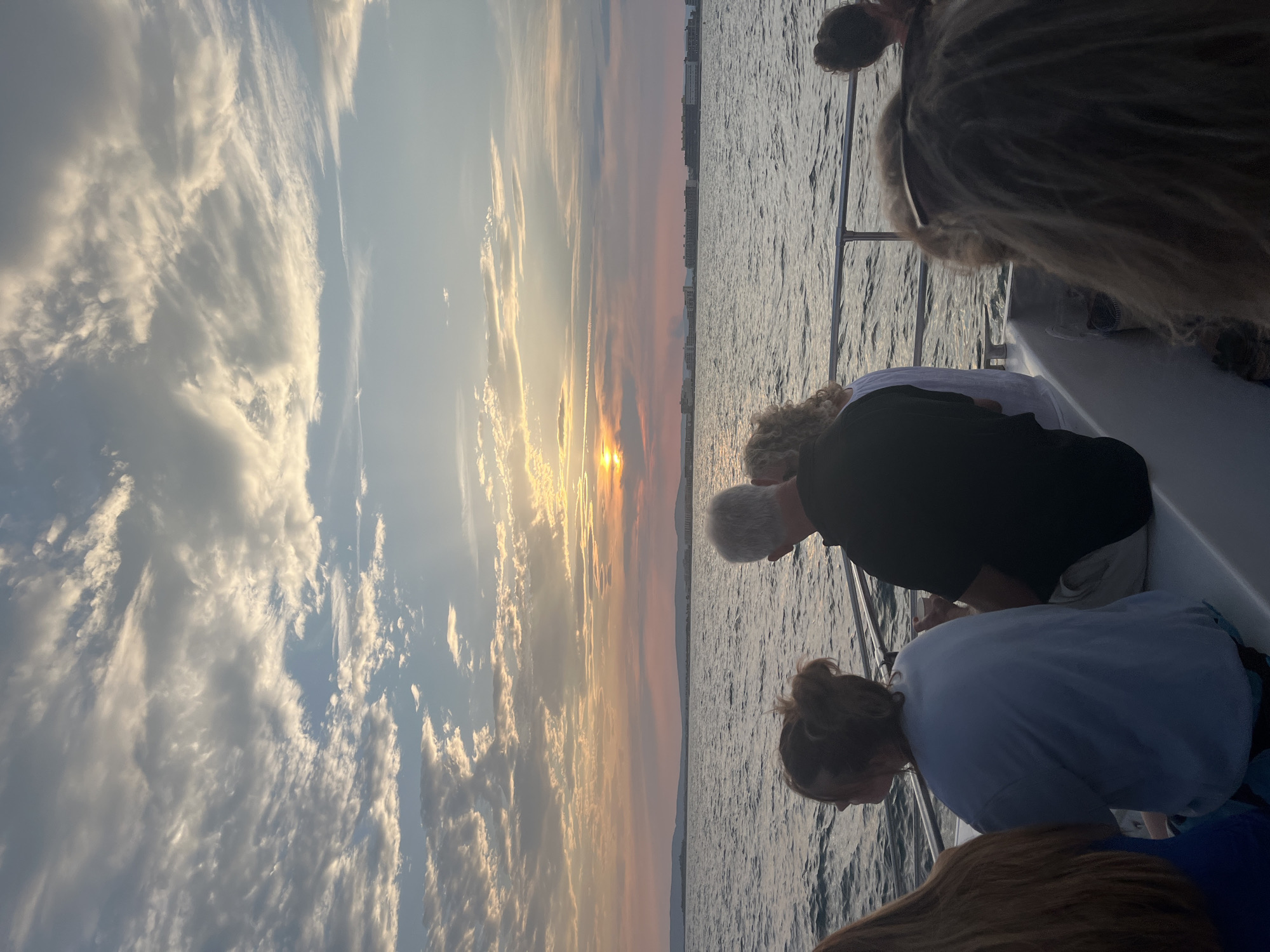 The image depicts a group of people on a boat, facing away from the camera, looking out at a sunset over a body of water. The sky is filled with dramatic clouds, illuminated by the setting sun, creating a warm, colorful glow. In the distance, a shoreline with buildings is visible. The water appears calm with gentle ripples. The people are casually dressed, suggesting a leisurely outing. The scene captures a peaceful moment at dusk, likely a tourist activity in Palavas-les-Flots, France, given the coastal setting.