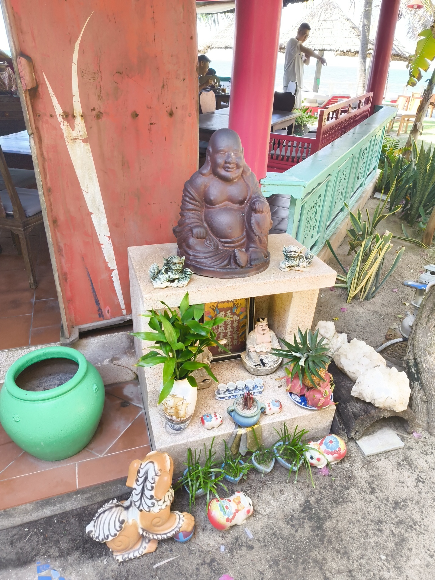 The image depicts an outdoor scene in Phu Thuy Ward, Vietnam, likely a garden area or patio of a restaurant or resort, given the presence of seating and structures suggestive of a dining or relaxation space. In the foreground, several decorative items are arranged on the ground and on a two-tiered stone pedestal.
Dominating the center of the pedestal is a dark brown, seated Buddha statue with a prominent belly and a cheerful expression. To its left, a small, intricate green dragon figurine rests on the upper tier of the pedestal. Below the Buddha statue, on the lower tier, is a small, seated Buddha figurine wearing a crown, placed in a decorative dish. Next to this, a white vase holds a green leafy plant. Further to the right on the lower tier, a small pile of incense sticks sits in a blue ceramic pot. A decorative plate holds a pineapple and two dragon fruits. Behind these items, several small white quartz-like rocks are scattered on the sand.
To the left of the pedestal is a large, bright green, circular ceramic pot. In the immediate foreground, on the concrete ground, are several decorative ceramic figurines of animals, including a dog-like creature with a patterned mane and tail, and two colorful, plump, rabbit-like figures. Small potted plants are also scattered among these figurines.
The background reveals a red, weathered wall on the left, with white diagonal markings. Behind the pedestal, a light blue and mint green railing with ornate carvings separates the foreground from a raised seating area. A man in a white t-shirt and glasses is visible in the background, standing near a red railing and pointing, seemingly interacting with someone out of frame. Further in the distance, tables, chairs, and thatched-roof umbrellas suggest an outdoor dining or seating area overlooking a body of water, likely the sea, under a bright sky. Palm trees and other greenery are visible throughout the scene. The ground is a mixture of concrete, sand, and patterned tiles.