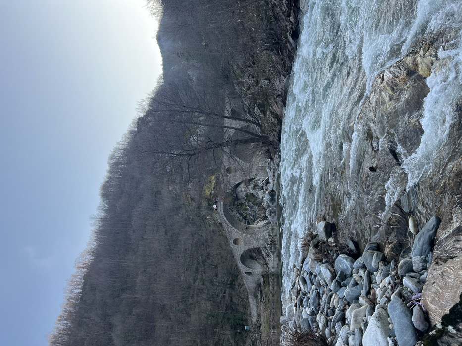 The image displays a natural landscape in Diadovtsi, Bulgaria, centered on a multi-arched stone bridge spanning a turbulent river. The river occupies the foreground and midground, exhibiting rapid, white-capped flow over visible rocks and a pebble-strewn left bank. The bridge, constructed from grey stone, features at least three visible arches and circular openings above the main arches. Two individuals are present on the highest segment of the bridge's leftmost visible arch; one appears to be seated or crouched, the other standing adjacent. The background is dominated by a steep hillside densely covered with bare deciduous trees, indicative of late autumn or winter conditions, under a clear, light blue sky.