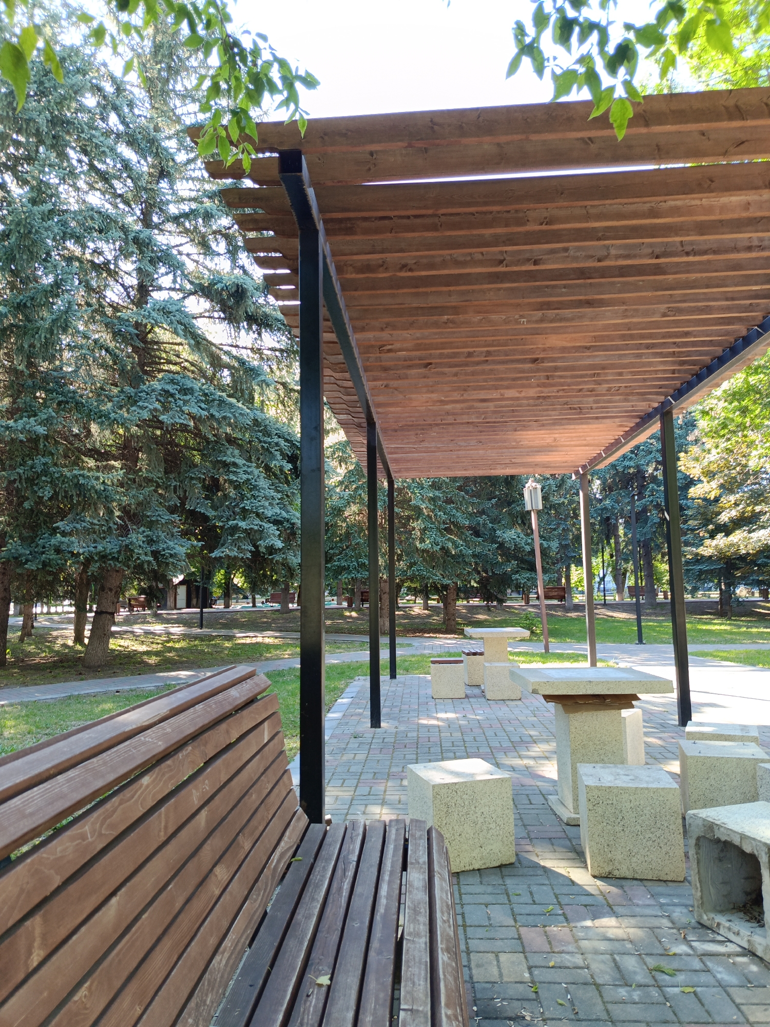 A wide-angle, eye-level outdoor shot captures a paved area in a park setting in Ust-Dzheguta, Russia. The foreground features a wooden park bench on the left, angled away from the camera. To the right and extending into the midground is a covered seating area with a wooden slatted roof supported by dark metal posts. Underneath the shelter, several concrete cube-shaped seats and at least one concrete table are arranged on the paved ground. The background is dominated by mature evergreen trees, some of which appear to be spruce or fir, casting dappled shadows on the ground. Sunlight filters through the trees and the slats of the roof, creating a bright and pleasant atmosphere. The park appears to be well-maintained, with paved walkways visible among the trees. No people are present in the scene.