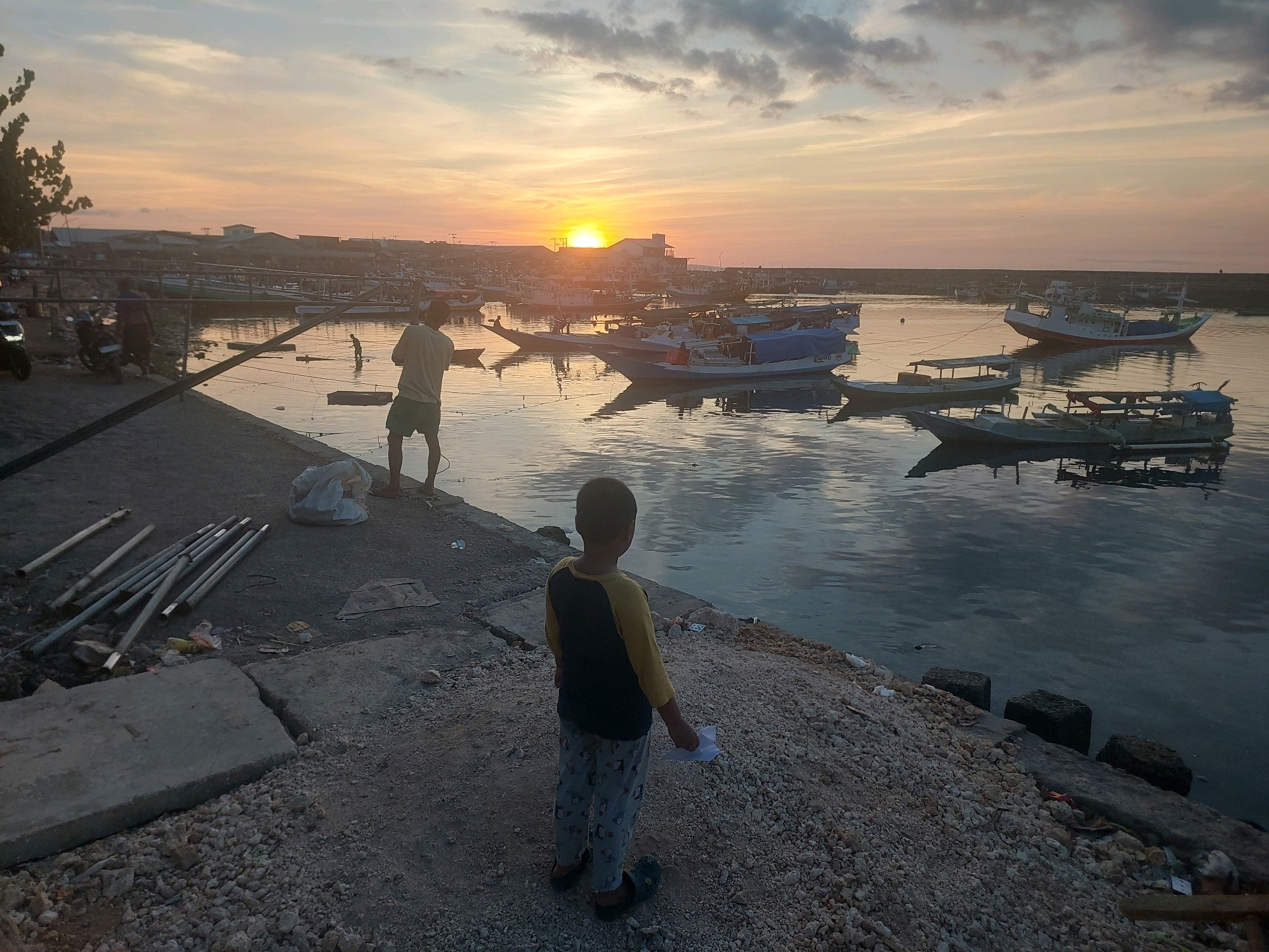 This image depicts a sunset scene at the Kupang harbor in Kupang, Indonesia. In the foreground, a young boy stands on a rocky embankment, facing away from the camera and towards the water. He is wearing a yellow and dark blue long-sleeved shirt and patterned pajama pants. He is holding a piece of white paper in his right hand. Behind him, a man in a light-colored shirt and shorts stands near the edge of a concrete pier, also facing the water. In the middle ground, numerous fishing boats of various sizes are docked and anchored in the calm harbor waters. The setting sun is visible on the horizon, casting a warm, orange glow across the sky and reflecting on the water. The background shows buildings and structures along the coastline, suggesting an urban or port environment. The scene is characterized by the soft light of dusk and the peaceful atmosphere of a harbor at the end of the day.
