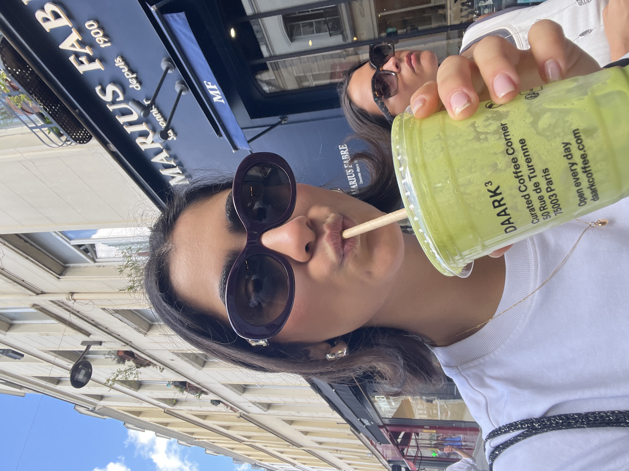 The image shows two women in Paris, France, posing for a selfie while one of them sips a green drink from "DAARK3 Curated Coffee Corner" at 50 Rue de Turenne, 75003 Paris. The drink's container displays the website "daarkcoffee.com" and states they are "Open every day." Behind the women, signage reads "MARIUS FABRE Savonnier depuis 1900". The sign also contains the label "MF". Building architecture and a partly sunny sky are visible in the background.