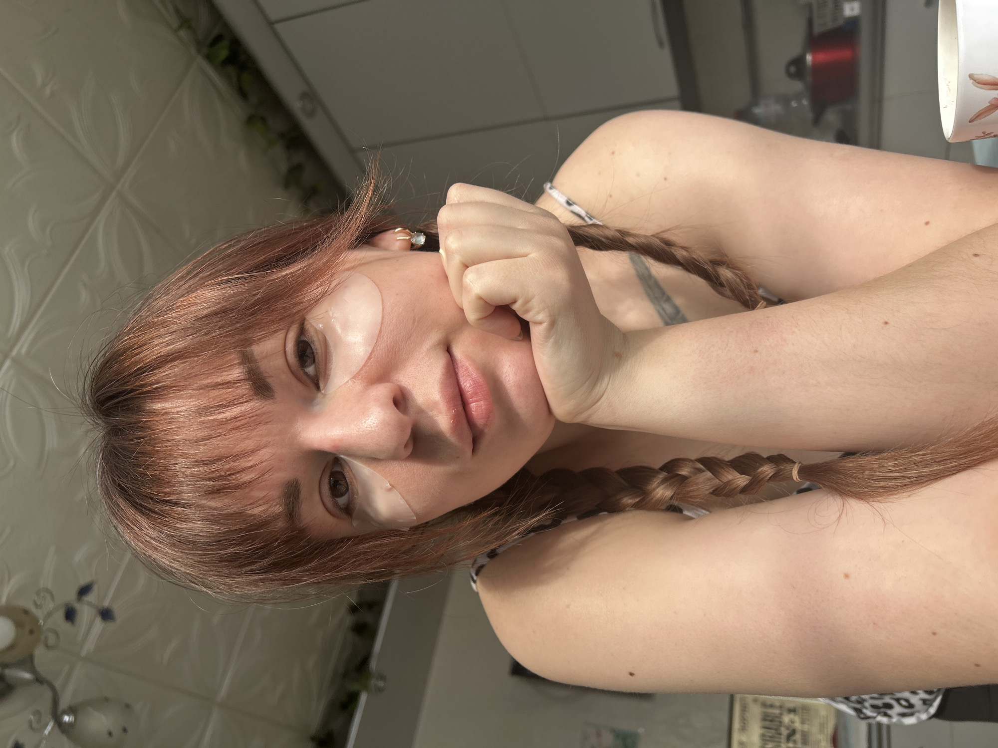 This image depicts a fair-skinned female subject with reddish-brown hair and bangs, styled with two visible braids extending over her shoulders. She is seated indoors, identified as a location within the city of Фоки, Russia. The subject is wearing transparent under-eye patches and gold-colored hoop earrings with light-colored gemstone drops. She is looking directly at the viewer with a neutral expression, resting her chin on her fisted right hand. A portion of a tattoo is visible on her left shoulder/upper arm. She is wearing a sleeveless top with a light-colored animal or abstract pattern. The background suggests a domestic kitchen or dining area, featuring a white textured ceiling with a light fixture, white overhead cabinets, and green foliage visible above them. On the right, a partially visible white mug with a patterned design, a red kettle, and a clear bottle are discernible on a counter surface.