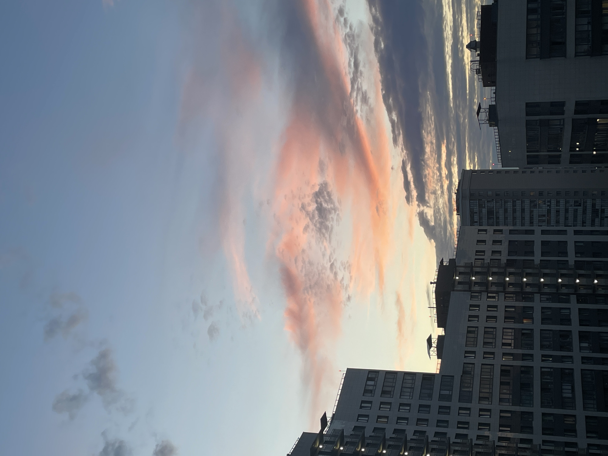 The image is a vertical, full-frame shot of a sky filled with pink and grey clouds, seen at sunset over a city. In the foreground, the upper sections of several modern apartment buildings are visible, with balconies and numerous windows. The buildings are dark grey and white. The sky transitions from a light blue at the top to warmer hues of pink and orange near the horizon, where the clouds are illuminated by the setting sun. Several small, dark grey clouds are scattered across the upper part of the sky.