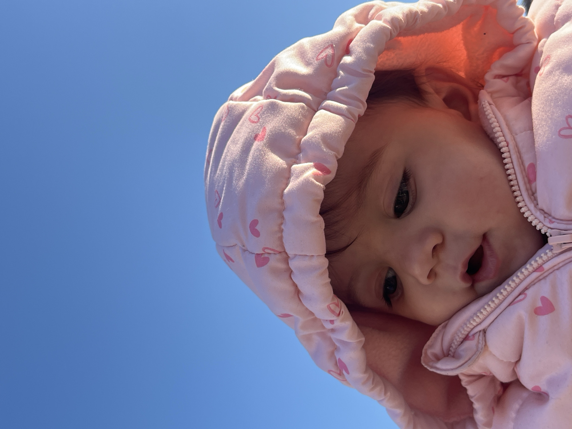 A baby with brown hair wearing a pink hooded jacket with hearts on it is looking down at the camera. The background is a bright blue sky. The location in the city of Elena, Bulgaria is not discernible from the image.