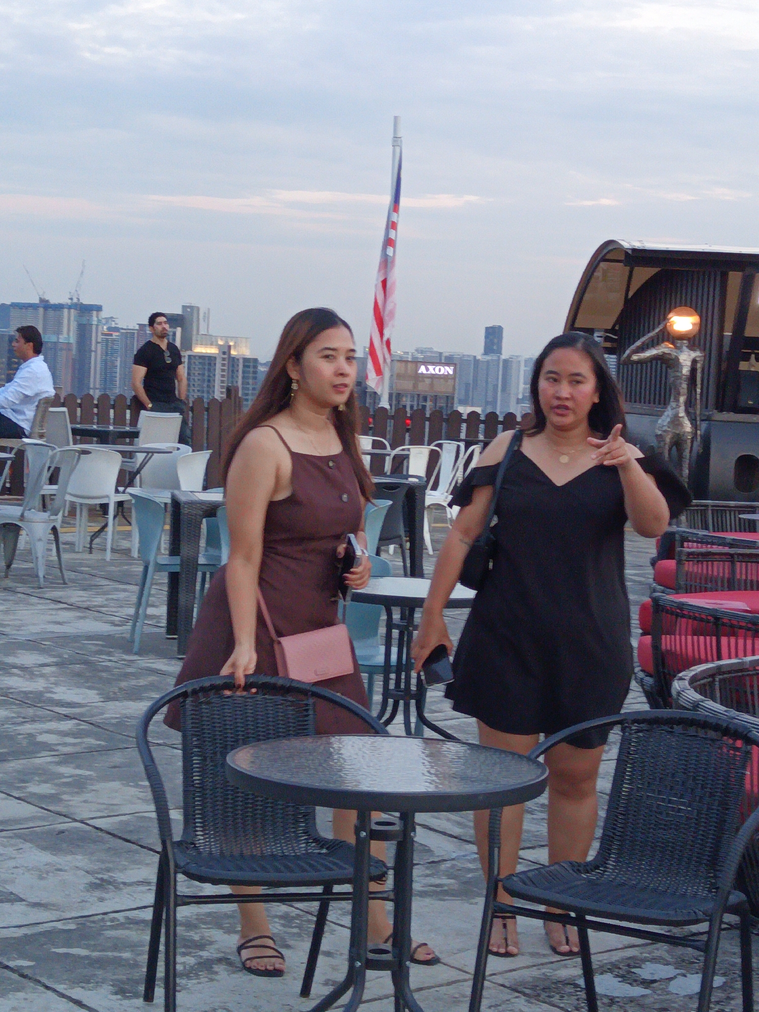 The image depicts a rooftop bar or restaurant in Kuala Lumpur, Malaysia, with a cityscape in the background. Two women are the central focus. The woman on the left wears a maroon sleeveless dress and carries a pink purse. The woman on the right is dressed in a black off-the-shoulder dress and points towards the viewer with her right hand. Several outdoor tables and chairs are visible, some with red cushions. An American flag is displayed on a pole to the left of the two women. The sky is overcast.