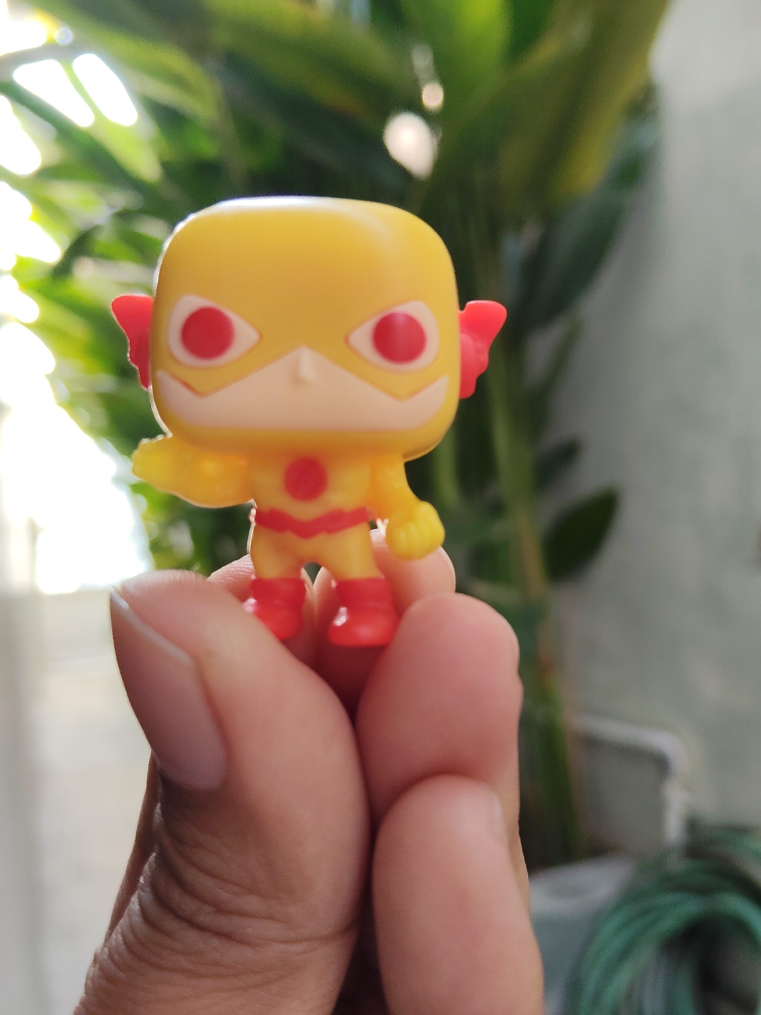 A yellow Funko Pop figure, resembling The Reverse-Flash from DC Comics, is held between two fingers of a person's left hand. The figure has red eyes, red ear wings, a red lightning bolt emblem on its chest, and red boots and gauntlets. The background is blurred and appears to be an outdoor setting with green foliage and sunlight filtering through. The image was taken in the city of Pemalang, Indonesia.