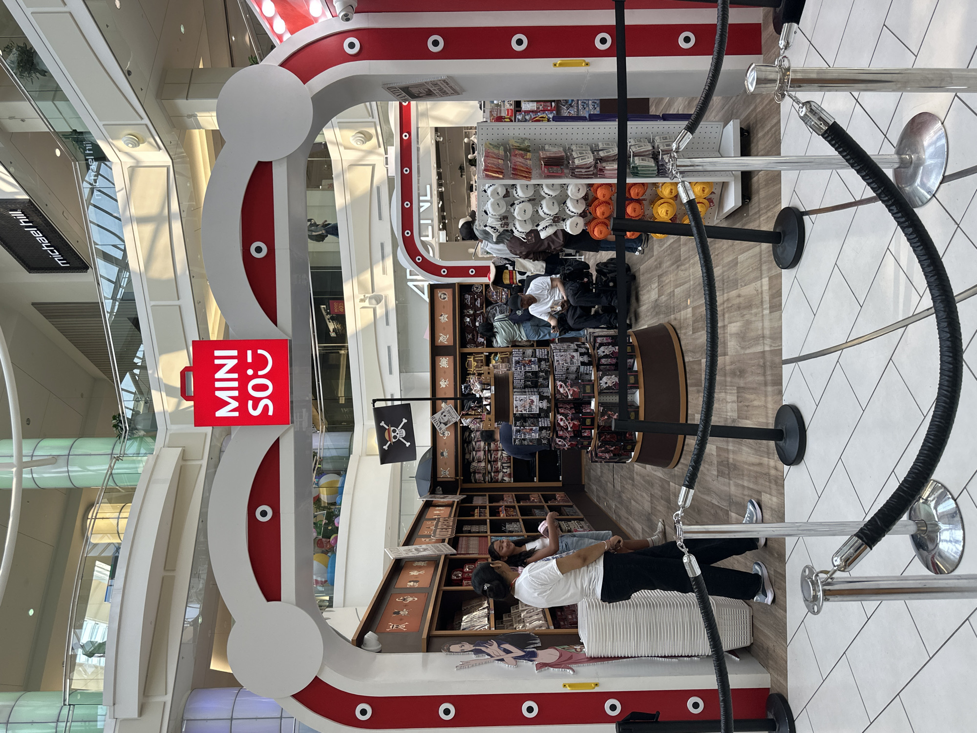 This is a full-shot, eye-level, outdoor photo taken of a retail kiosk designed to resemble a whimsical, stylized whale, with a large red sign displaying "MINISO" at the top of the arch. The kiosk is situated within a shopping mall, as evidenced by the polished floor tiles, the multi-level architecture with balconies, and signage for businesses like "michael hill." Inside the kiosk, shelves are stocked with merchandise, including what appears to be anime or manga-related collectibles, such as figurines and posters. A black pirate flag with a skull and crossbones is displayed. Several individuals are visible: two women are standing near the entrance to the kiosk, one appearing to be a customer and the other an employee. A man in a wheelchair is seated inside the kiosk, browsing merchandise. Other customers are also visible, some interacting with the products. Black velvet ropes on stanchions cord off the entrance to the kiosk, suggesting a controlled flow of customers. The overall environment suggests a lively shopping atmosphere. The location is identified as Metropolis at Metrotown in Burnaby, Canada.