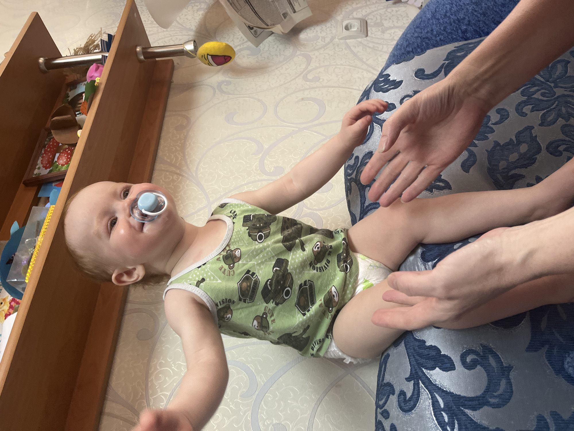 A baby wearing a green tank top with cartoon animal images and a diaper is lying on a blue patterned cushion. A pacifier is in the baby's mouth. Two hands are extended toward the baby. The scene appears to be indoors, with a wooden shelf or bookcase in the background containing toys and other objects. The wall is decorated with a light-colored patterned wallpaper. There is a yellow toy hanging above the baby. The location is Chaikovsky, Russia.