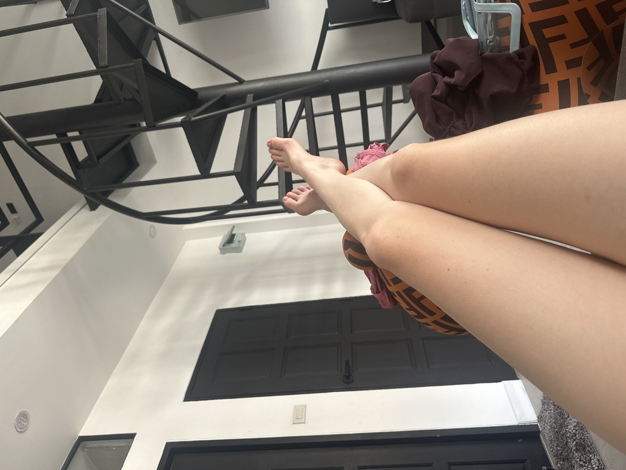 A low-angle shot looking up shows a person's bare legs crossed in the foreground, resting on a pillow. The location appears to be inside a building in Baan Chaweng, Thailand, suggested by architectural elements. The background includes a staircase, a wall with a black window or door frame, and possibly an air conditioning unit. The focus is on the legs and the setting.