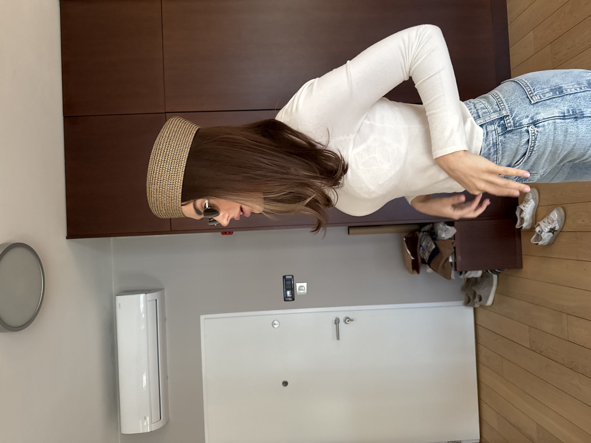 The image shows a woman with long brown hair wearing a straw headband and sunglasses. She is wearing a white long-sleeved shirt and light blue jeans. She is standing in front of a dark brown cabinet. To the left of the cabinet is a white wall with a round light fixture and an air conditioner unit. Below the cabinet is a white door with a handle and a peephole. On the floor to the right of the door is a wooden floor and several pairs of shoes. The location is an interior space of unknown location in Unknown, Unknown.