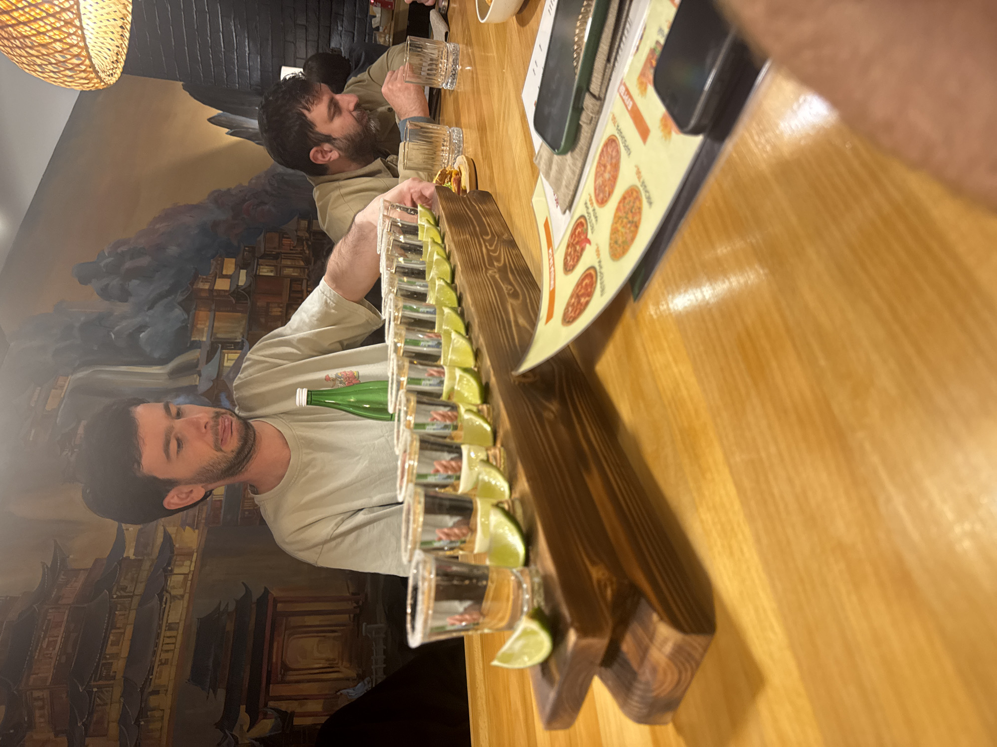 The image depicts a social gathering at a restaurant in Sokhumi, Abkhazia. Two men are seated at a wooden table. One man is facing the camera, wearing a light-colored shirt, while the other is facing away. A wooden tray containing multiple shot glasses filled with a clear liquid and garnished with lime wedges is on the table, suggesting a round of tequila shots. A green bottle is also on the table. A menu rests on the table near the shot glasses. In the background, a wall is adorned with a mural depicting a cityscape. A woven lampshade is visible overhead.