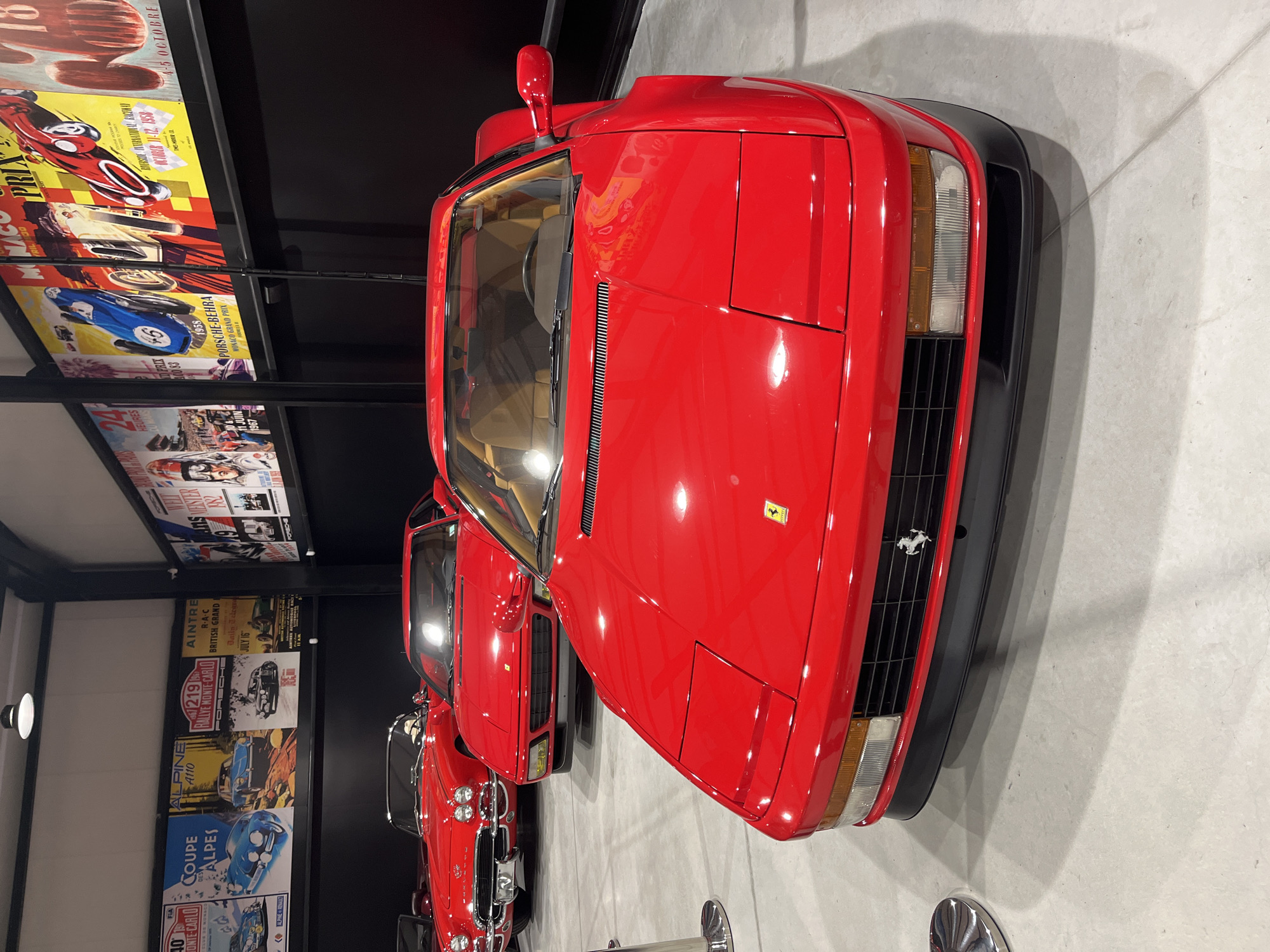 A high-angle, eye-level shot depicts a row of classic red cars, likely Ferraris, in what appears to be a museum or showroom. The first car dominates the frame with its front end prominently displayed. Behind it, other red cars are partially visible. The environment includes posters of vintage races and cars displayed on a black wall to the left. The scene appears to be indoors with bright lighting. There is no indication of this being in Kapatovo, Bulgaria.