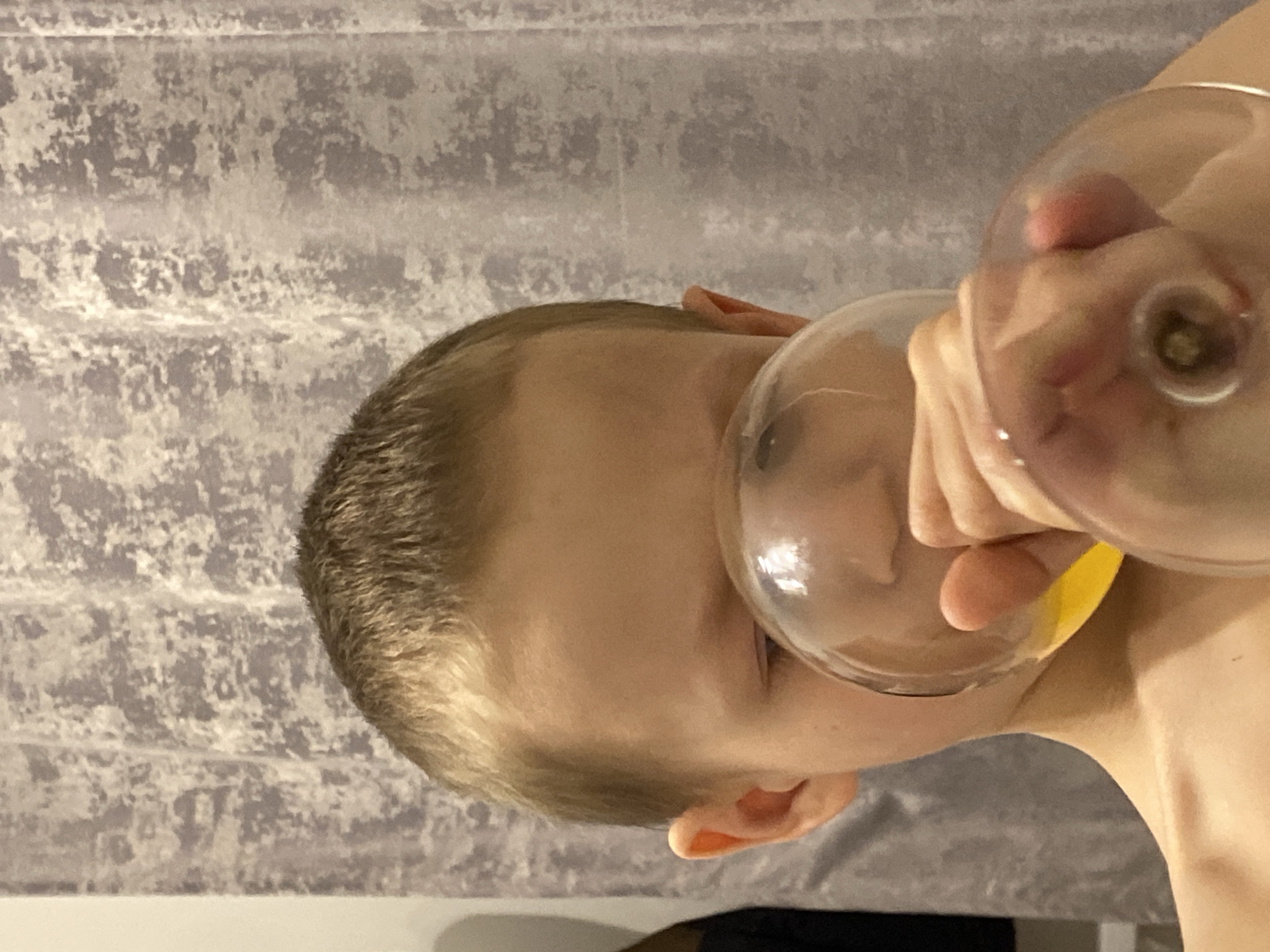 A fair-skinned child is holding two glass objects, possibly wine glasses, to their face. The glasses appear to be partially filled with liquid. The child is indoors, and the background is a textured grey surface. The image does not contain information to determine the location in Saint Petersburg, Russia.