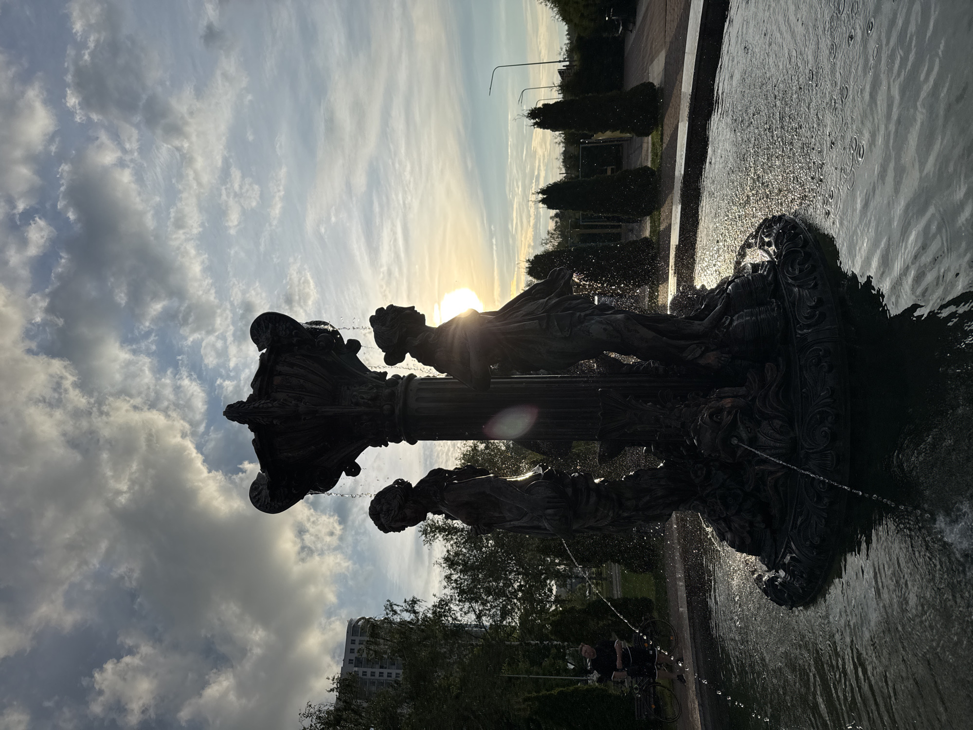The image displays the "Mermaids" (or "Nymphs") fountain situated in the Svisloch River Embankment Park, near Victory Park on Pobediteley Avenue, Minsk, Belarus. The scene is captured during the late afternoon or early evening, evidenced by the low sun creating a strong backlight effect.
The central subject is a large, ornate bronze fountain, featuring two primary female classical figures standing on a lower tier, with an elaborate upper basin from which water cascades. Both statues are rendered in silhouette against the bright sky, with the sun's disc visible as a prominent flare to the right of the left statue's chest. Water sprays and drips from various points of the fountain into a circular basin, where sunlight creates sparkling highlights and ripples on the dark surface.
The sky above is partially cloudy, presenting a mix of darker, textured cumulus clouds and lighter, more ethereal clouds, interspersed with patches of blue. To the left of the fountain, a tall, multi-story, light-colored modern residential building is visible through a dense line of trees, including both deciduous and coniferous varieties. In the mid-ground on the left, partially obscured by a tree and the fountain's spray, a person is seen standing near or riding a bicycle. To the right of the fountain, a paved pedestrian pathway recedes into the background, flanked by a row of neatly trimmed, dark green conical coniferous trees. Further in the distance on the right, two vertical streetlights or utility poles are discernible against the lighter horizon.