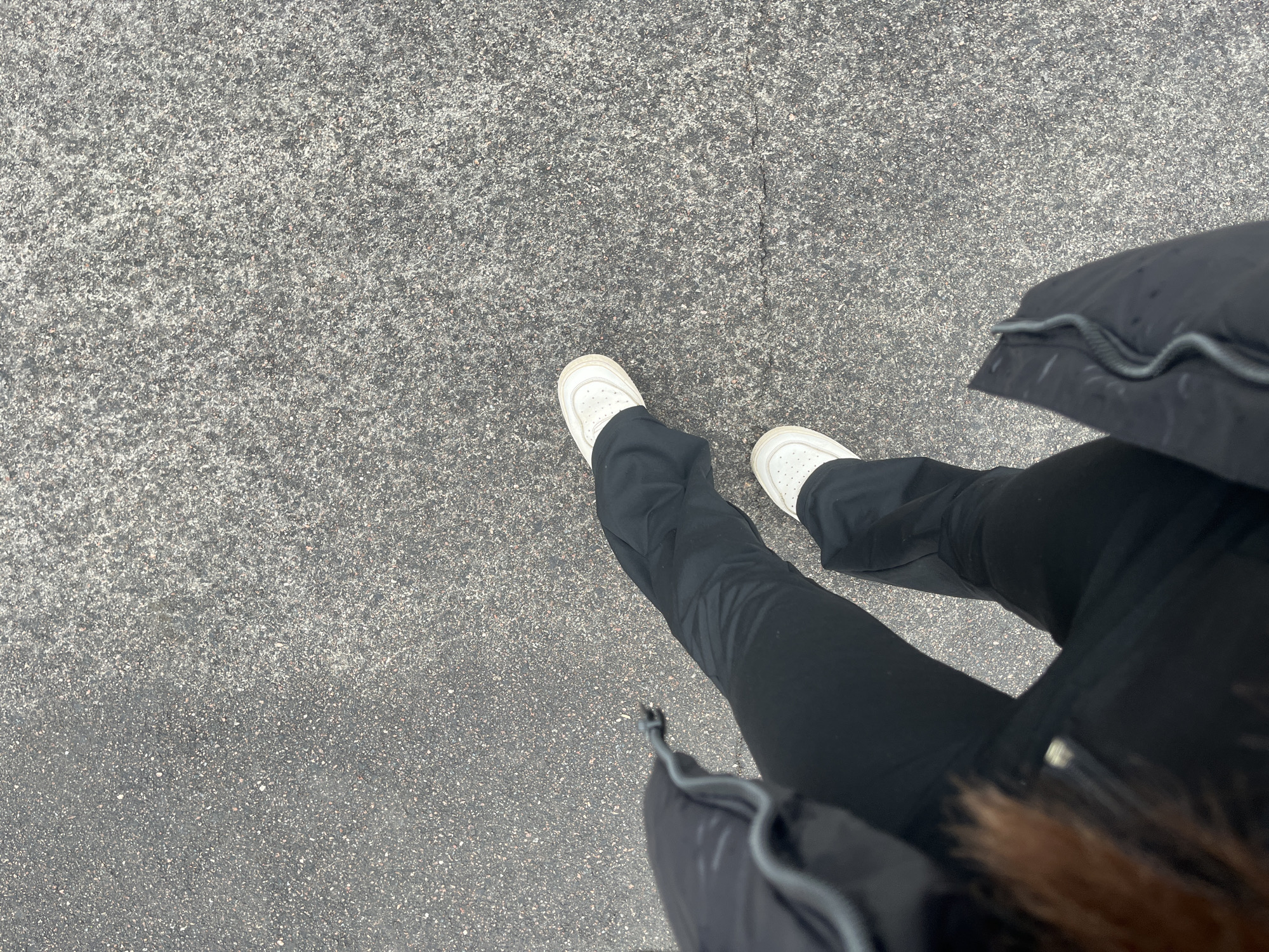 The image shows a first-person perspective of a person standing on a gray asphalt surface. The person is wearing black pants and white sneakers. Part of a black winter coat with fur trim is visible on the right side of the frame. The asphalt appears to be a sidewalk or pavement. The location can't be confirmed as Saint Petersburg, Russia based on this image.