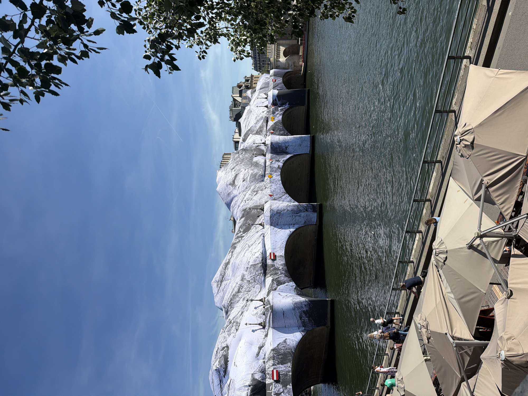 This image shows the Pont Neuf in Paris, France, wrapped in silver fabric. The bridge spans the Seine River. On the right side of the frame, along the riverbank, there are several beige umbrellas shading an outdoor dining area where people are sitting. The sky is blue with a few wispy clouds and some foliage is visible in the top left corner of the image.