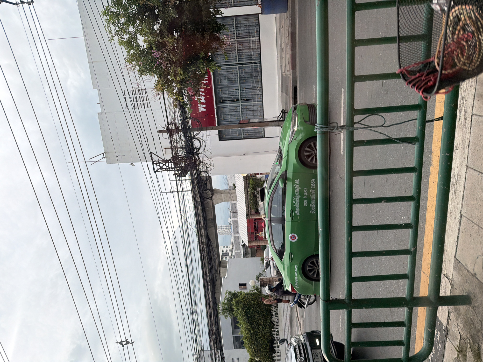 The image depicts a street scene in Bangkok, Thailand, with a prominent green taxi driving on the right side of the road. The taxi has Thai script and numbers on its side, including "2022 4567" and "081-701-0701". In the foreground, a green metal railing with a yellow stripe runs along the sidewalk. A black mesh basket, containing what appears to be woven items and red rope, is attached to the railing.
To the left of the taxi, a person wearing a helmet rides a motorcycle, also on the road. Further to the left, a black car is partially visible. The background features several buildings, including a white building with windows and a red sign with Thai writing. Overhead, a dense network of electrical wires crisscrosses the sky, a common sight in Bangkok. The sky is overcast, suggesting a cloudy day. Trees with pinkish flowers are visible on the right side of the image. The overall scene captures a typical urban environment in Bangkok, with traffic, infrastructure, and urban foliage.