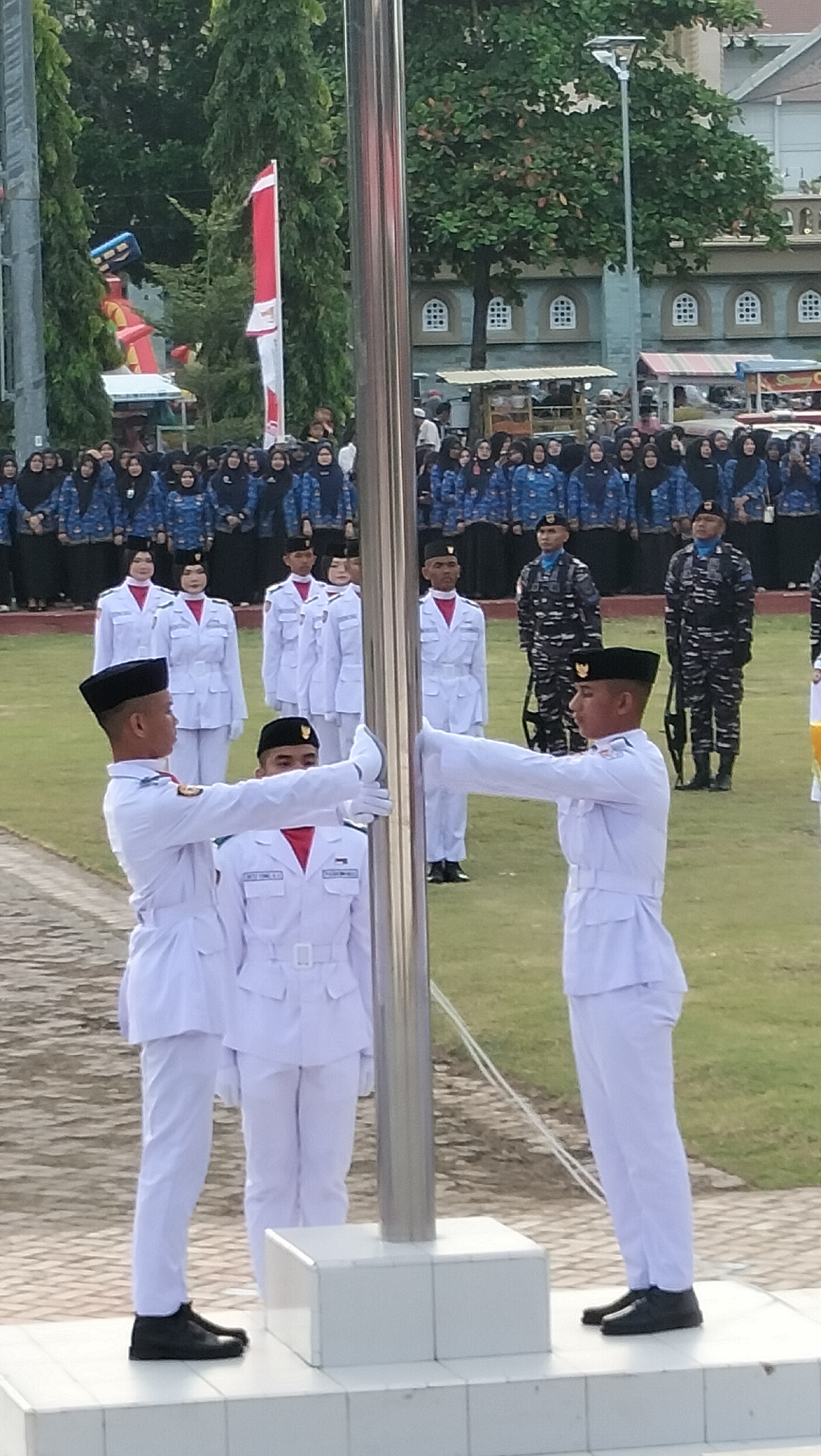 In Lhokseumawe, Indonesia, a flag-raising ceremony is depicted. Two young men in white military-style uniforms and black caps are positioned in the foreground, holding a metal flagpole. Both individuals have their right hands on the pole, appearing to be actively involved in its movement. They are standing on a white elevated platform. Behind them, a group of individuals in similar white uniforms are also present. In the background, a larger assembly of people, predominantly women wearing blue patterned tops and headscarves, are observing the ceremony. Behind them, buildings with distinctive arched windows and trees are visible, suggesting an institutional or educational setting within the city. The overall scene conveys a sense of formality and organized event.