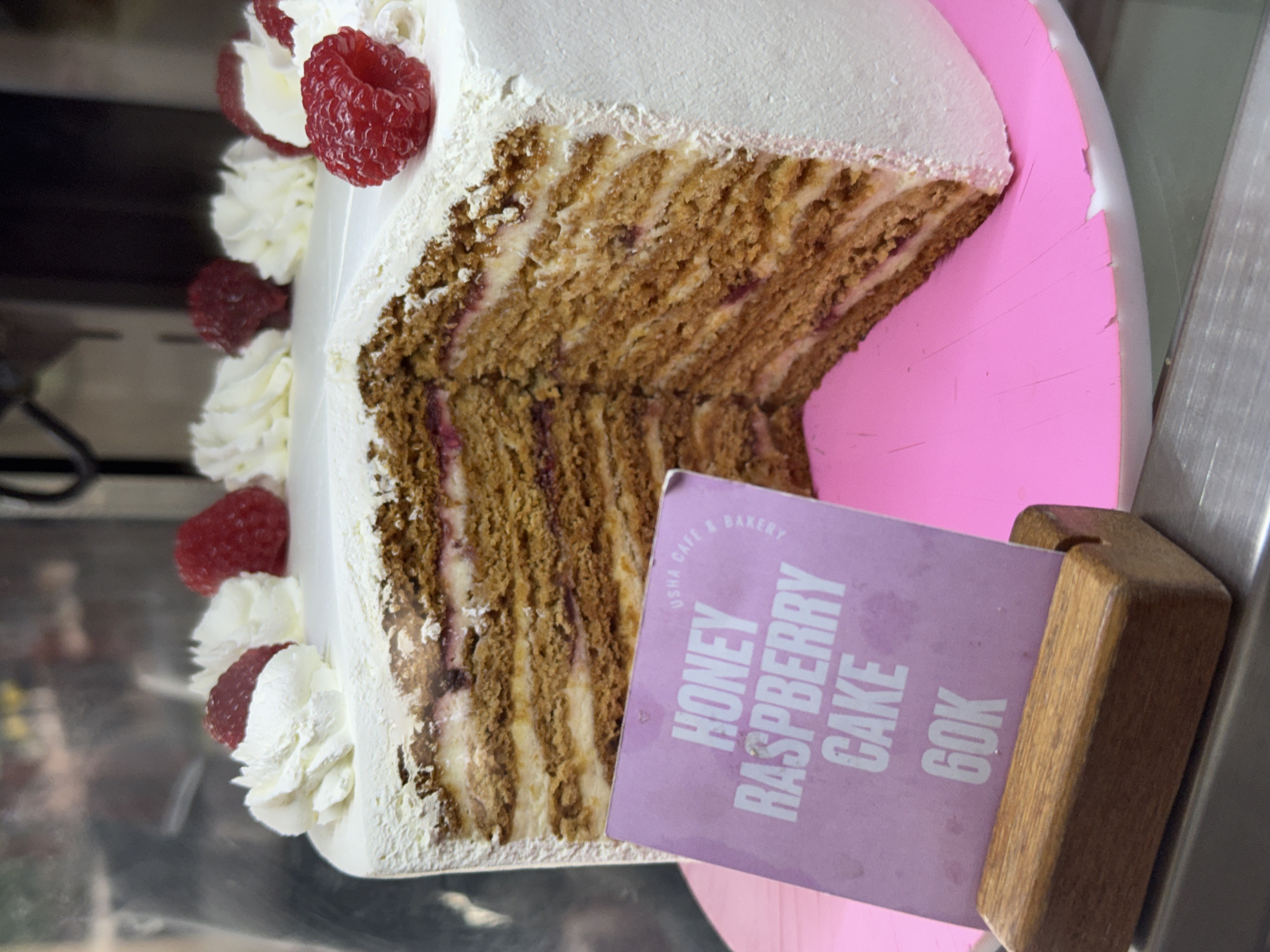 A close-up, side-view shot showcases a slice of honey raspberry cake. The cake has multiple layers of light brown sponge cake and white cream, interspersed with visible raspberry filling. It's topped with white frosting and garnished with fresh raspberries and dollops of whipped cream. The slice is presented on a pink cake board, which appears slightly damaged at the edge. Leaning against the cake board is a light purple card with text printed in white. The text reads: "USHA CAFE & BAKERY", "HONEY RASPBERRY CAKE", and "60K". The cake slice is positioned in what appears to be a display case, with reflections on a metallic surface in the background. The lighting suggests an indoor setting, likely a cafe or bakery. There are no people visible in the image, and the focus is entirely on the cake and its presentation. The overall impression is one of a delicious, freshly prepared dessert ready for sale.