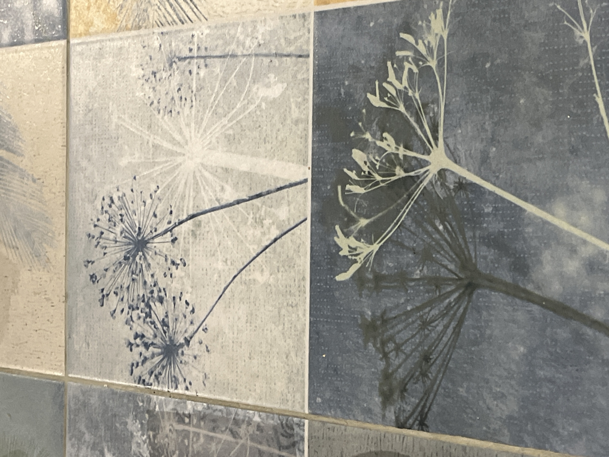 The image displays a close-up, high-angle shot of four decorative tiles arranged in a grid. The top two tiles feature a light blue and off-white abstract design resembling dandelion seeds in various stages of dispersal against a textured background. The bottom two tiles depict a dark blue background with a contrasting light-colored silhouette of a dill-like plant and a dark silhouette of a similar plant. The tiles are arranged on a flat surface, possibly a wall or floor, with visible grout lines between them. No people or specific location details are discernible.
