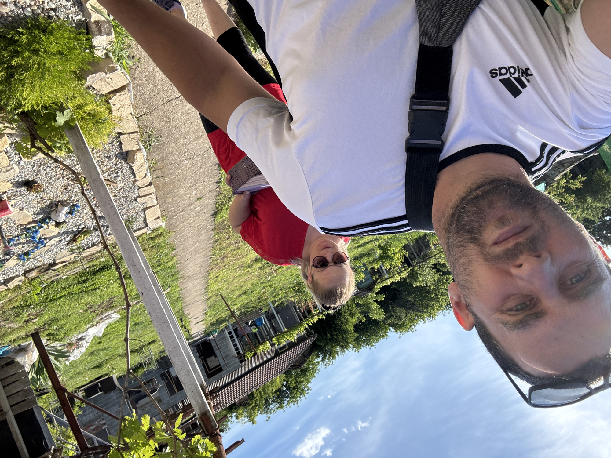 A slightly tilted, eye-level selfie photograph shows two people in Gorna Lipnitsa, Bulgaria. The foreground features a man with short dark hair and a beard, wearing glasses and an Adidas shirt, and a strap from a bag crosses his torso. Behind him, a woman with blonde hair, wearing sunglasses and a red shirt, stands on a path. The background includes vegetation, a blue sky with clouds, and buildings. A concrete post and some plant growth are in the immediate left foreground.