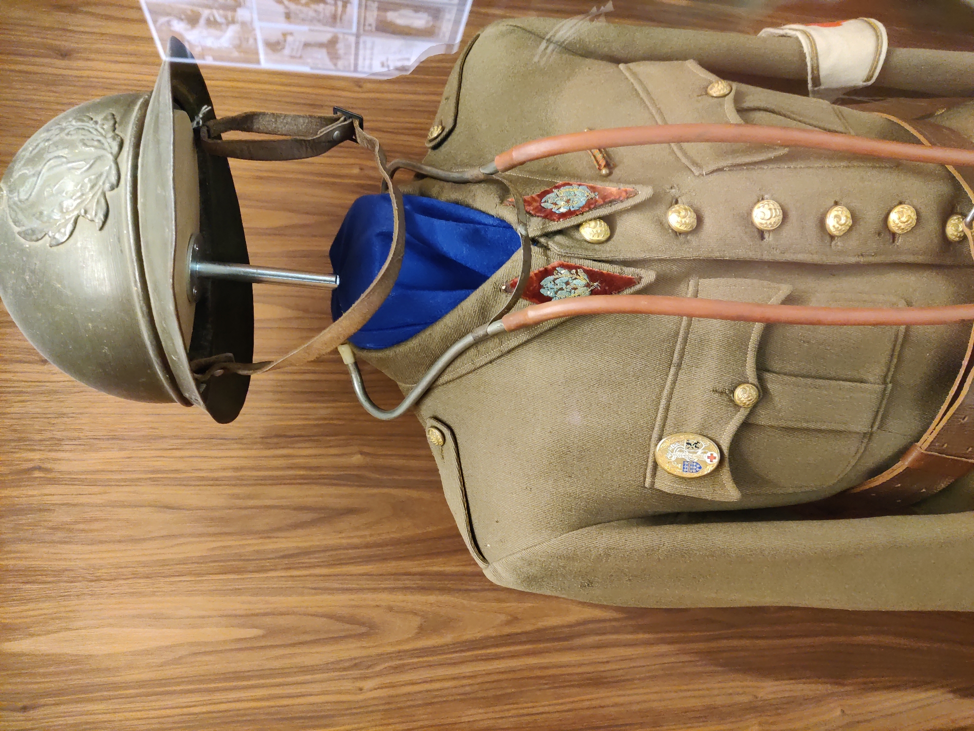 This is a medium close-up, eye-level shot of a vintage military medical uniform displayed in a museum. The uniform is khaki-colored and includes a helmet, jacket, and belt. The jacket has several gold buttons, two breast pockets with flap closures, and epaulets. A blue scarf or cravat is visible at the neckline, and a stethoscope is draped around the neck and over the jacket. The helmet is olive green with a metallic emblem on the front. A white armband with a red cross is visible on the right sleeve. The background is a wooden paneled wall, and to the right of the uniform, a display of historical photographs is partially visible. The overall impression is one of historical preservation and display. The location is specified as the Dunkirk Museum in Dunkirk, France.