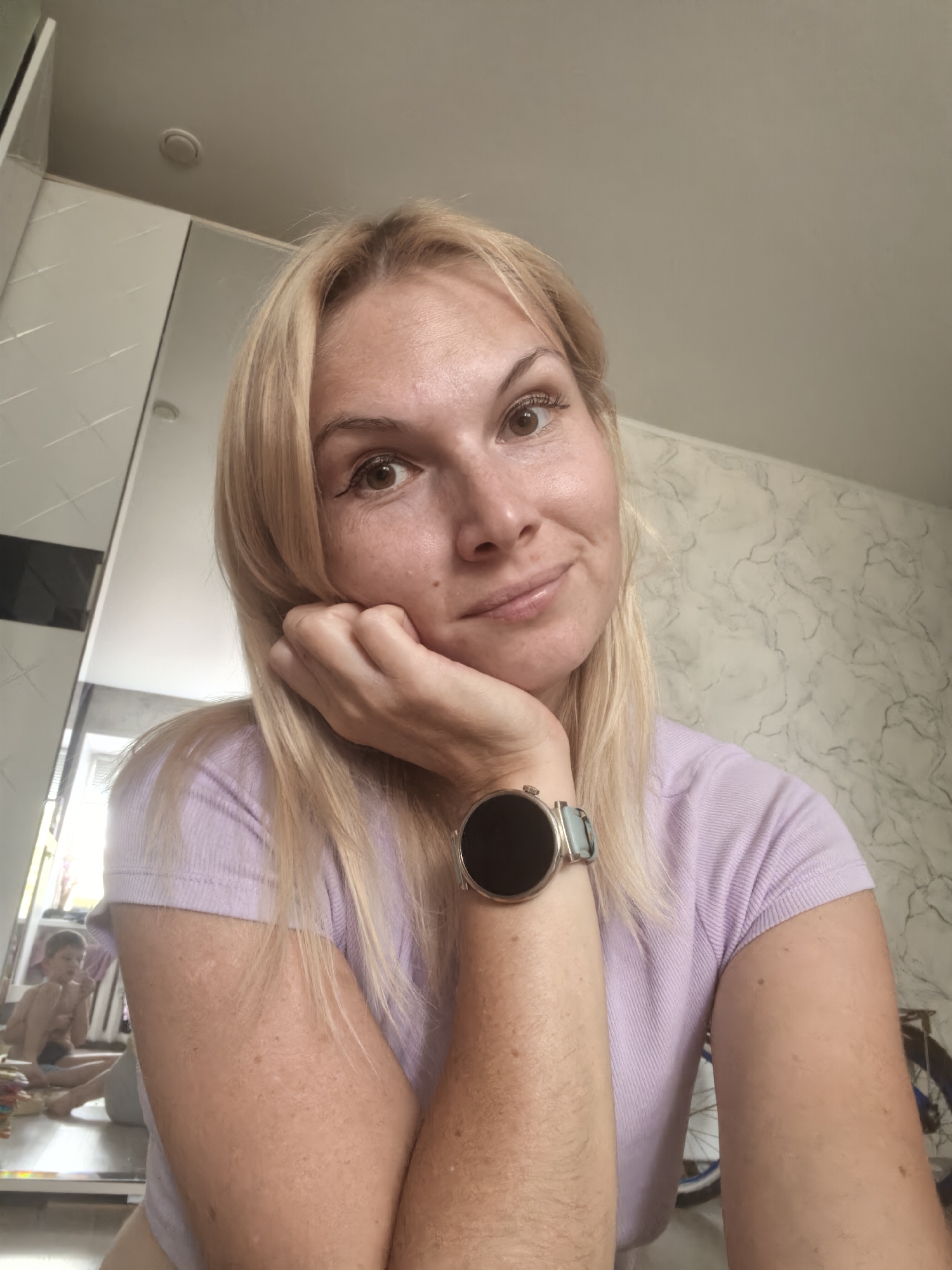 The image is a selfie of a blonde woman with her left hand resting on her chin. She is wearing a light purple t-shirt and a smart watch with a light blue band on her left wrist. The background includes a wall with a marble-like pattern and a large mirror on the left side, which reflects a young person in the background. The overall setting appears to be indoors, possibly a room in a residential building. No specific location in Sterlitamak, Russia, can be identified from the image.