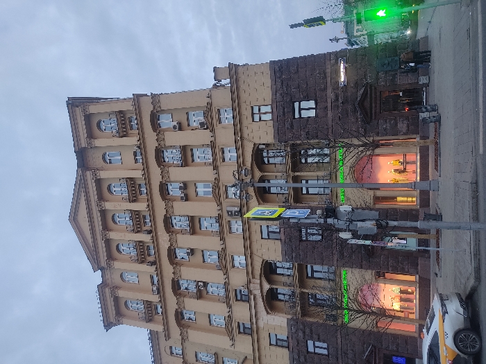 The image depicts a street scene in Moscow, Russia, focusing on a multi-story building. The building's facade is divided into two distinct sections: the left side is a lighter, yellow-tan color with classical architectural details, including columns and arched windows, and numerous air conditioning units are affixed to the exterior. The right side of the building is constructed with darker, roughly textured stone. The ground floor houses a shop or commercial space, visible through large display windows. A sign with green lettering is above. A pedestrian crossing sign is attached to a metal pole in the foreground. Traffic signals are visible at the top of the frame, showing a green light. A vehicle, possibly a taxi, is parked on the right edge of the frame. People are standing in front of the building entrance. The sky is overcast. The scene suggests an urban setting with a mix of historical architecture and modern commercial activity.