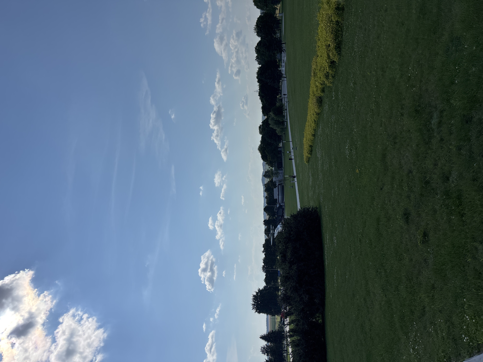 The image depicts a bright, sunny day with a clear blue sky scattered with fluffy white clouds. The foreground is dominated by a lush, green grassy field. In the middle ground, a row of vibrant yellow-green bushes forms a distinct line. Beyond the bushes, there's a paved pathway where several people are walking, appearing as small figures in the distance. Further back, a dense line of dark green trees stretches across the horizon. In the far distance, several buildings and vehicles, including what appears to be a large white structure resembling a boat or exhibition hall, are visible. The scene suggests an outdoor public space, possibly a park or museum grounds, with a sense of open space and clear weather. The location is identified as the Ozyaritska-Slabadski rural council in Belarus.