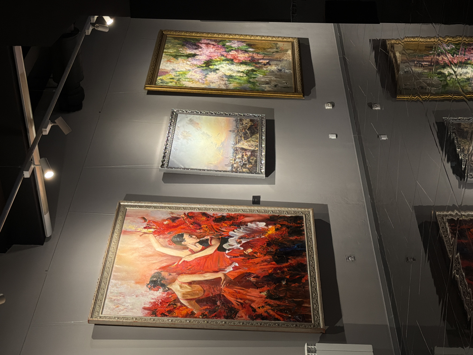The image depicts a section of an art gallery or exhibition space. Three framed paintings are prominently displayed on a dark gray wall. The painting on the left is large and features two female figures in vibrant red dresses, appearing to be dancing flamenco. The central painting is smaller and shows a cityscape at sunset. The painting on the right is vertical and depicts a colorful bouquet of flowers. The floor is a highly reflective dark surface, possibly polished stone or tile, which mirrors the paintings and the lighting above. Track lighting with adjustable spotlights is mounted on the ceiling, illuminating the artwork. A white radiator is visible on the far left of the wall, and electrical outlets are present on the wall between the paintings. The overall ambiance suggests a contemporary and well-lit exhibition space. The reference to Saint Petersburg, Russia, is not evident from the visual information in the image itself.