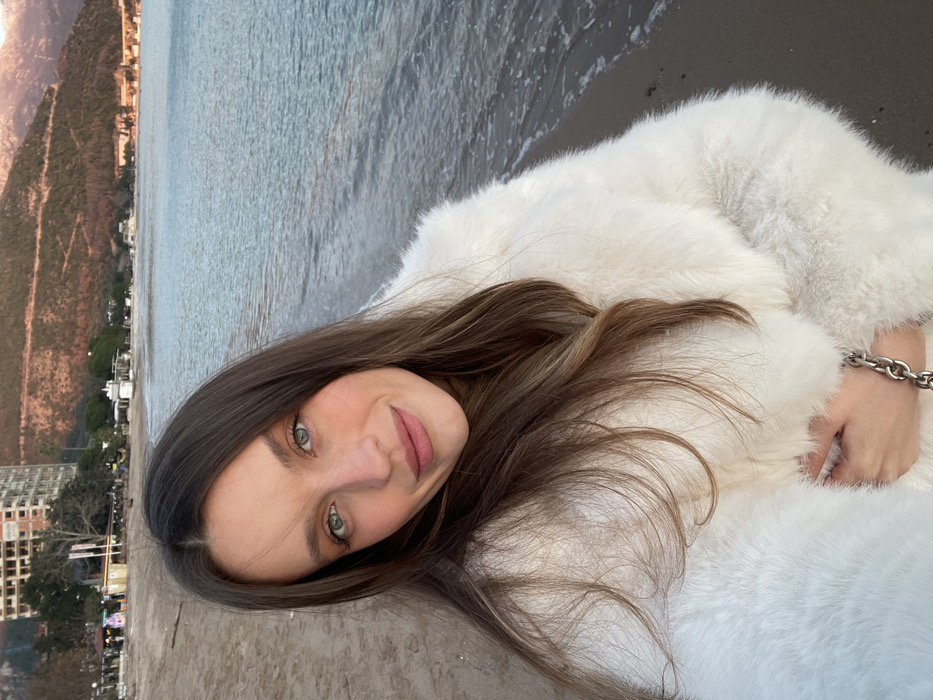 This portrait features a young woman lying on her side, gazing directly at the viewer with a calm and gentle expression. Her long, dark brown hair, highlighted with lighter streaks, frames her face and spreads around her shoulders. She has striking blue-green eyes and a soft, natural lip color. She is dressed in a plush white faux fur coat, and a thick silver-colored chain bracelet adorns her left wrist.
She appears to be resting on a dark, sandy or pebbled beach. Behind her, an expanse of calm, grayish-blue sea stretches out, meeting a distant coastline. This coastline is characterized by reddish-brown mountains descending to the water's edge, where numerous white and light-colored buildings line the shore. The soft, even lighting suggests either an overcast day or the golden hour of dusk or dawn. The overall ambiance is serene. The image, while presented in a vertical orientation, appears to have been taken in landscape mode and then rotated 90 degrees clockwise, causing the woman's head to be sharply tilted and the horizon to run diagonally across the frame. There is no discernible text. The mountainous coastal scenery is indicative of a location like Budva, Montenegro.