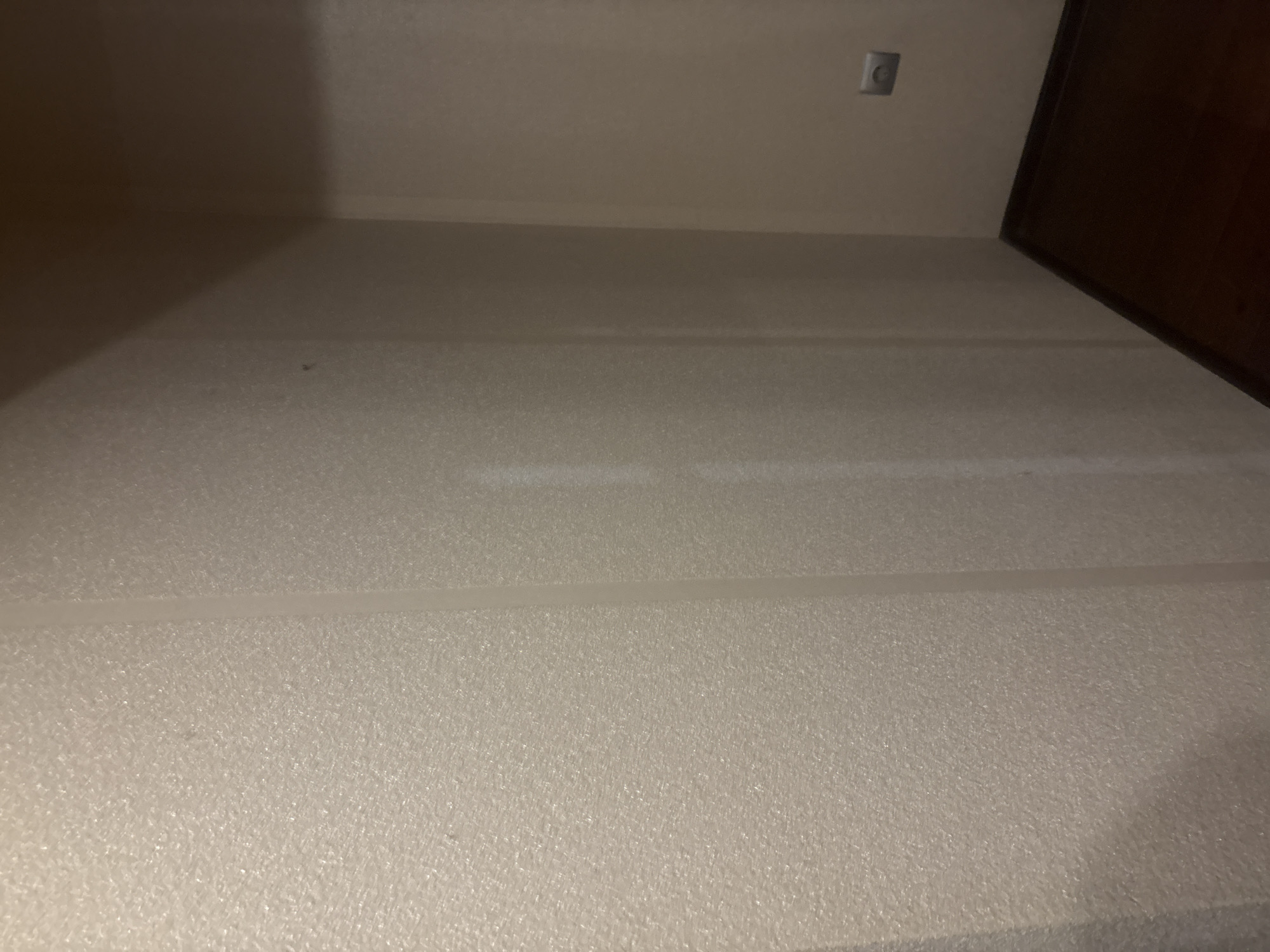 The image shows an interior view of a corner in a room with textured, light-colored wallpaper on the left wall and a darker, smoother wall on the right. The floor is dark wood. A light-colored vertical seam is visible on the wallpapered wall, suggesting a wallpaper seam. On the right wall, there is a gray electrical outlet with a circular indentation. The lighting suggests an indoor environment, possibly dimly lit. The location in the city of Orekhovka, Russia, is not explicitly indicated by any visual cues in the image itself.