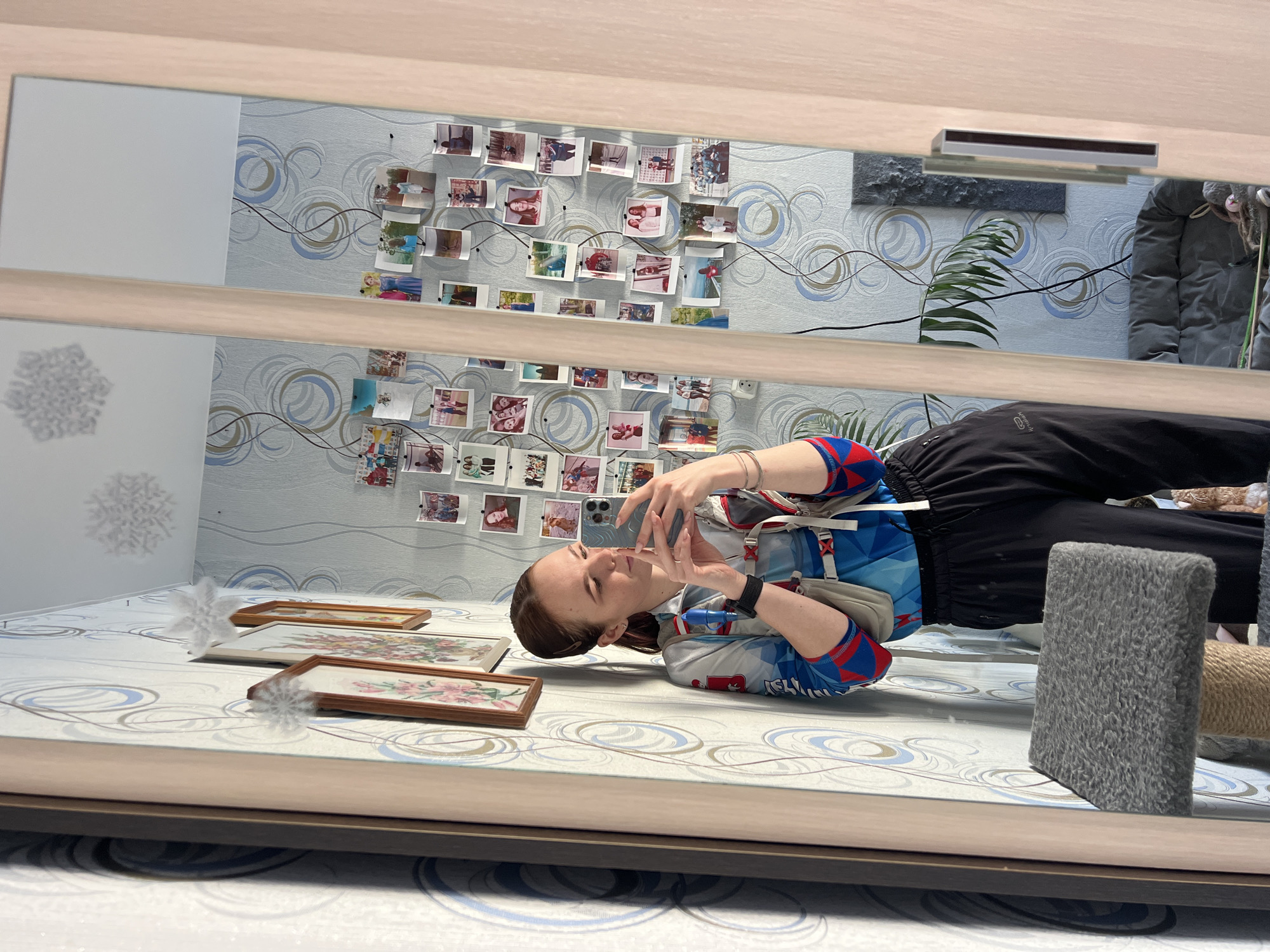 The image depicts an indoor scene in Chaikovsky, Russia, where a female individual is captured taking a selfie in a large mirror. The woman, with her brown hair pulled back, is wearing a blue, red, and white athletic long-sleeve top with geometric patterns, a light-colored hydration vest, and black athletic pants. She holds a light blue/grey smartphone with a multi-lens camera in both hands, positioned to reflect herself. A black digital watch is visible on her left wrist, and a silver bracelet is on her right.
The background directly behind the individual features light blue/grey wallpaper adorned with a pattern of concentric circles in blue, grey, and gold/brown tones. To the immediate left of the mirror, two framed artworks with floral designs hang on the wall, beneath two white, fluffy snowflake decorations. To the right of the mirror, numerous small, rectangular photographs are pinned directly to the wallpaper, connected by dark, thin strings, forming a collage.
In the lower-left foreground of the image, a grey, textured cat scratcher is partially visible. The mirror's reflection reveals further elements of the room: a houseplant with long green leaves, a dark grey pet bed with a small plush bunny toy on the floor, and a rectangular, textured grey wall hanging. Natural light appears to enter from the left, casting a dark shadow of a window or door frame onto the patterned wallpaper.