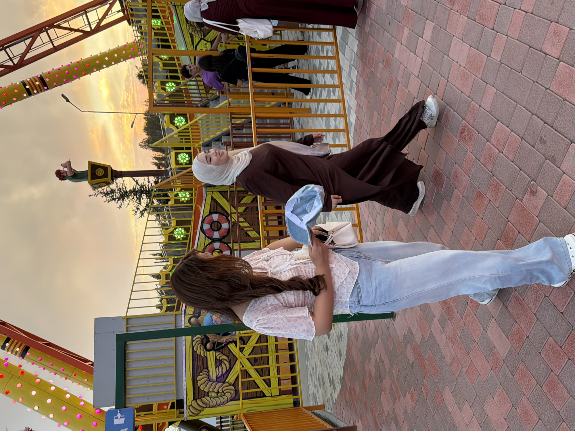 In an amusement park setting with a pirate ship ride visible in the background, two women are walking on a brick pathway. The woman in the foreground on the left is wearing a light-colored, floral print blouse and light blue wide-leg jeans. She has long, brown hair and is holding a light blue hat in her right hand and a small white purse in her left. To her right, another woman is walking in the opposite direction. She is wearing a long, dark brown abaya and a light beige hijab. She is also holding a light blue hat. The sky in the background displays a sunset with orange and yellow hues. The park appears to be open as other people are visible in the background, some near the ride entrance.