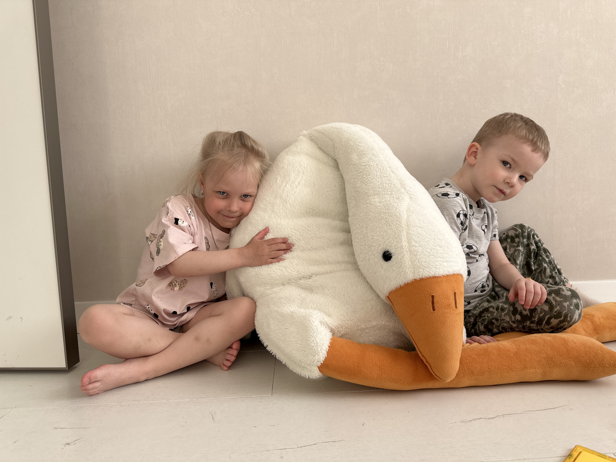 Two young children, a girl and a boy, are seated on the floor next to a large stuffed goose. The girl is blonde with her hair tied up, wearing a pink patterned pajama set, and she is hugging the goose. The boy has short brown hair and is wearing a gray t-shirt with soccer balls and camouflage pants. The goose is predominantly white with an orange beak and neck. The floor is light-colored, possibly wood or tile, and the background wall is a light beige color. The location is implied to be in Прикамский, Russia, but there are no visual cues to confirm the city.