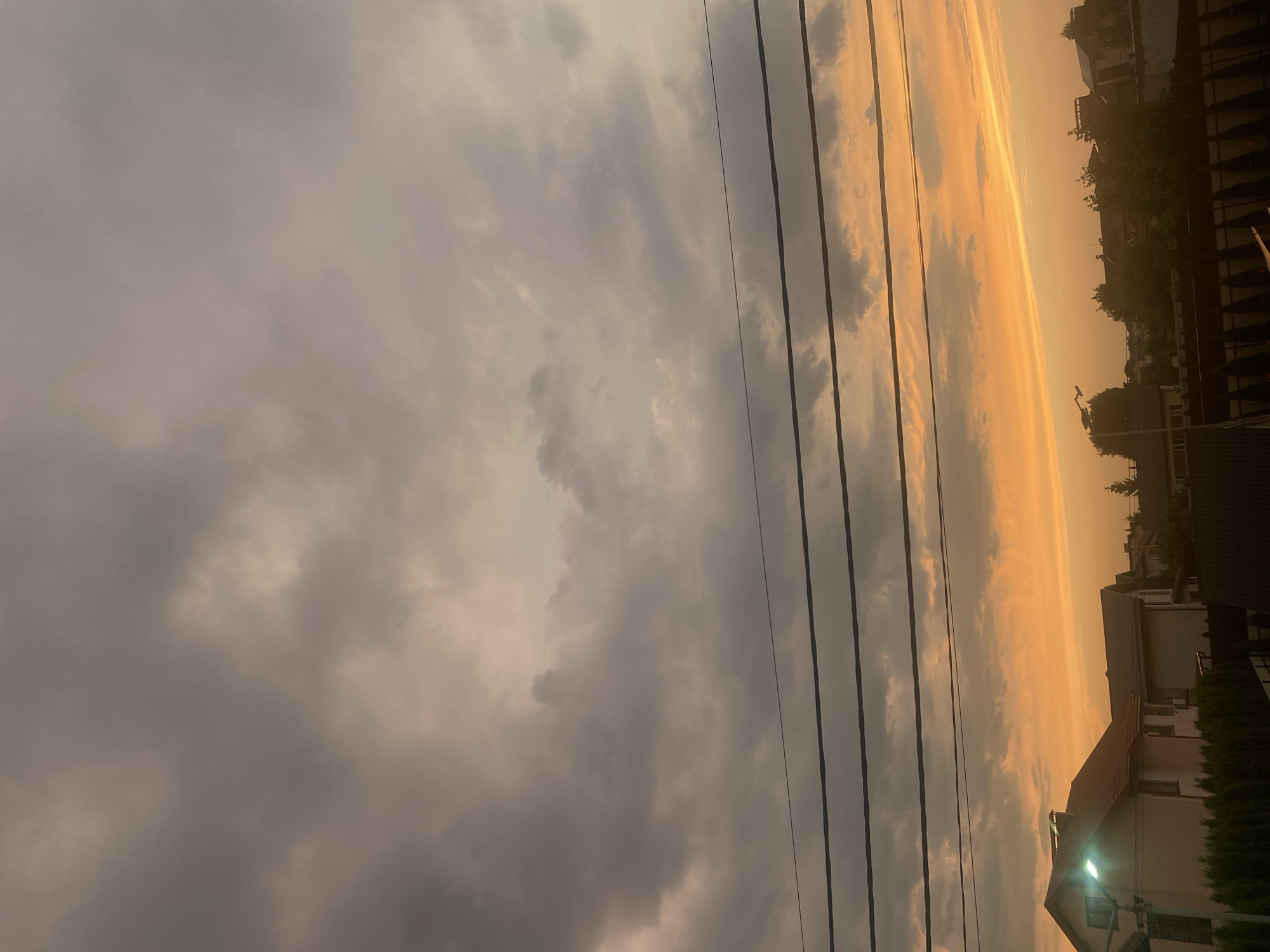 The image depicts a sky filled with heavy, gray clouds, with a sliver of bright orange and yellow on the horizon, indicating a sunset. Power lines are strung horizontally across the frame. Below the sky, the rooftops of houses, some with visible windows and one with a light on, are visible, along with trees and a fence. The setting appears to be a residential area in Novi Petrivtsi, Ukraine. There are no people or animals visible in the image.