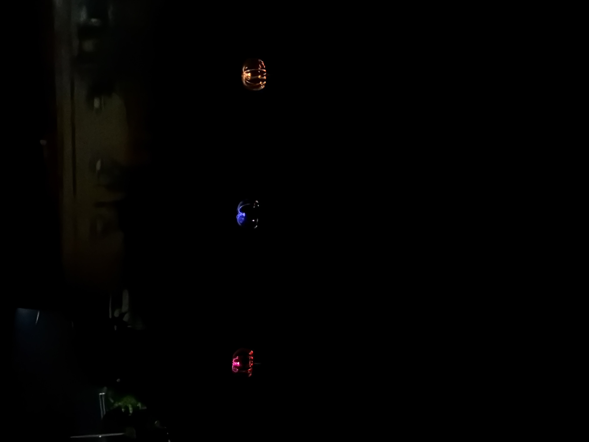 The image displays an extremely dark nocturnal outdoor scene in Akkuzovo, Russia, dominated by three horizontally aligned illuminated decorative objects. On the far left, a red and pink glowing light, possibly abstract or floral in design, is visible. A centrally placed blue and purple glowing light, similar in form, occupies the middle ground. To the right, an orange glowing object distinctly shaped like a carved pumpkin or jack-o'-lantern is present. The background is severely underexposed, rendering most details indistinct; however, faint, blurry outlines suggest distant structures or buildings and some dark foliage on the upper left. No individuals are present, and no actions or interactions are discernible. The environment appears to be an open outdoor space at night.