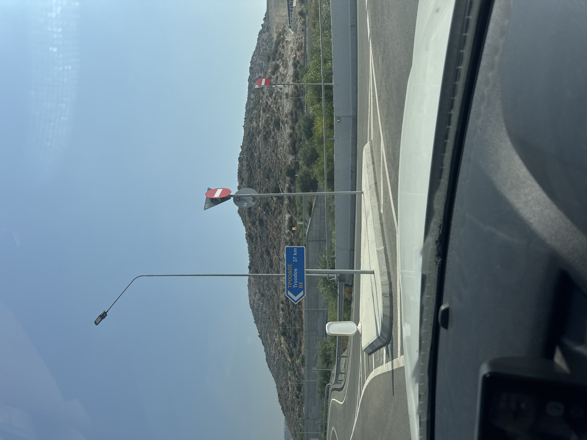 The image shows a street scene in Limassol, Cyprus, near a highway interchange. In the foreground, the dashboard of a car is visible. The road curves to the left, with white lane markings. A street lamp stands on the right side of the road. A blue directional sign points to the right, indicating "Troodos 37 km" and "B8". There are two "no entry" signs visible on poles. In the background, a barren, rocky hill rises, with some sparse vegetation. A concrete barrier separates the road from the hill. The sky is clear and blue. The overall impression is of a sunny, arid environment.