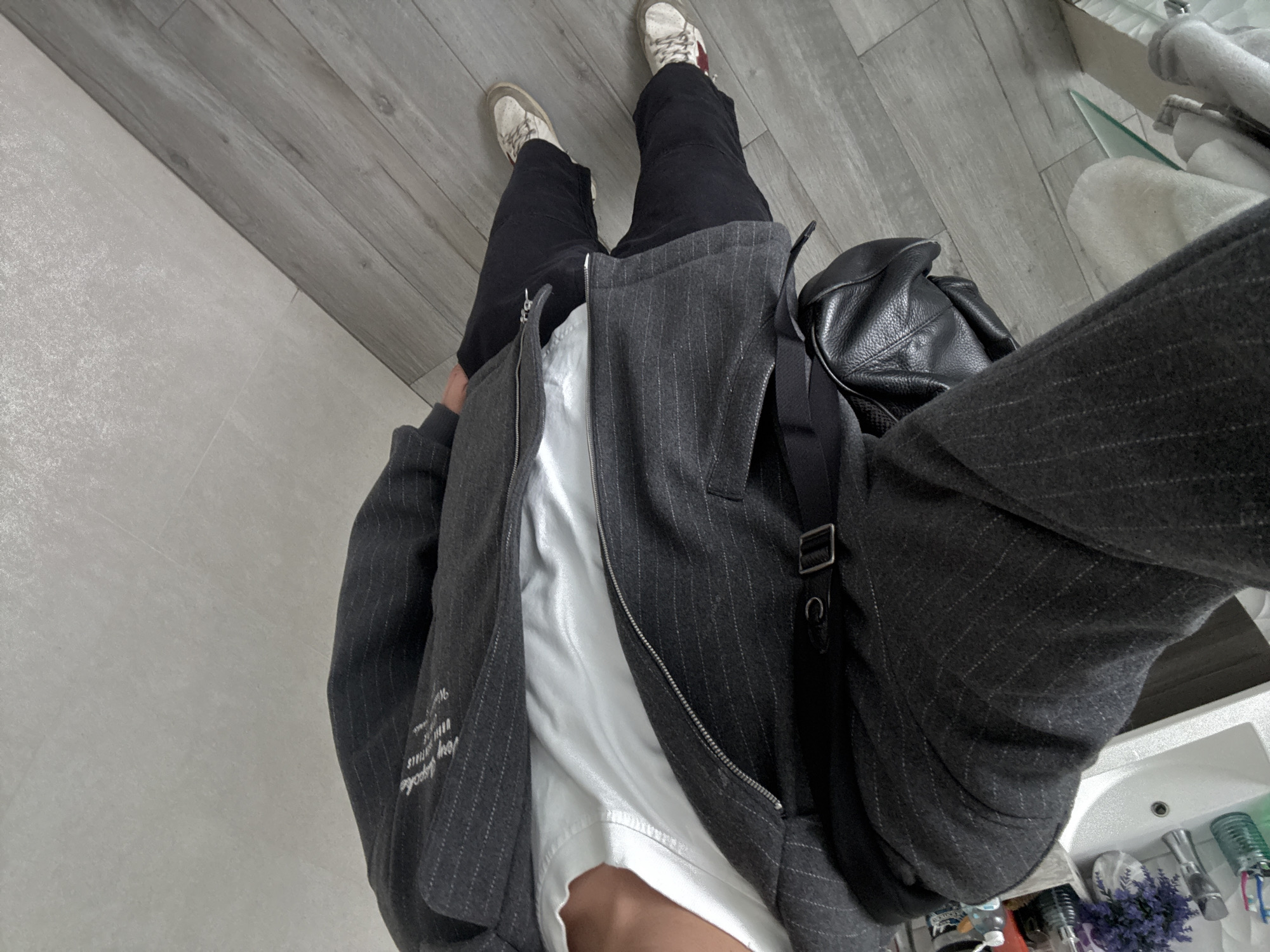 The image shows a person wearing a dark pinstripe jacket over a white t-shirt, black pants, and white sneakers. The person is carrying a black backpack and is standing in what appears to be a bathroom with a sink, a countertop with toiletries, and a tiled floor. The background features light-colored textured walls and wooden flooring. The image is taken from a first-person perspective, showing the person's legs and torso. The location is identified as being in the city of San Bernardino Tlaxcalancingo, Mexico.
