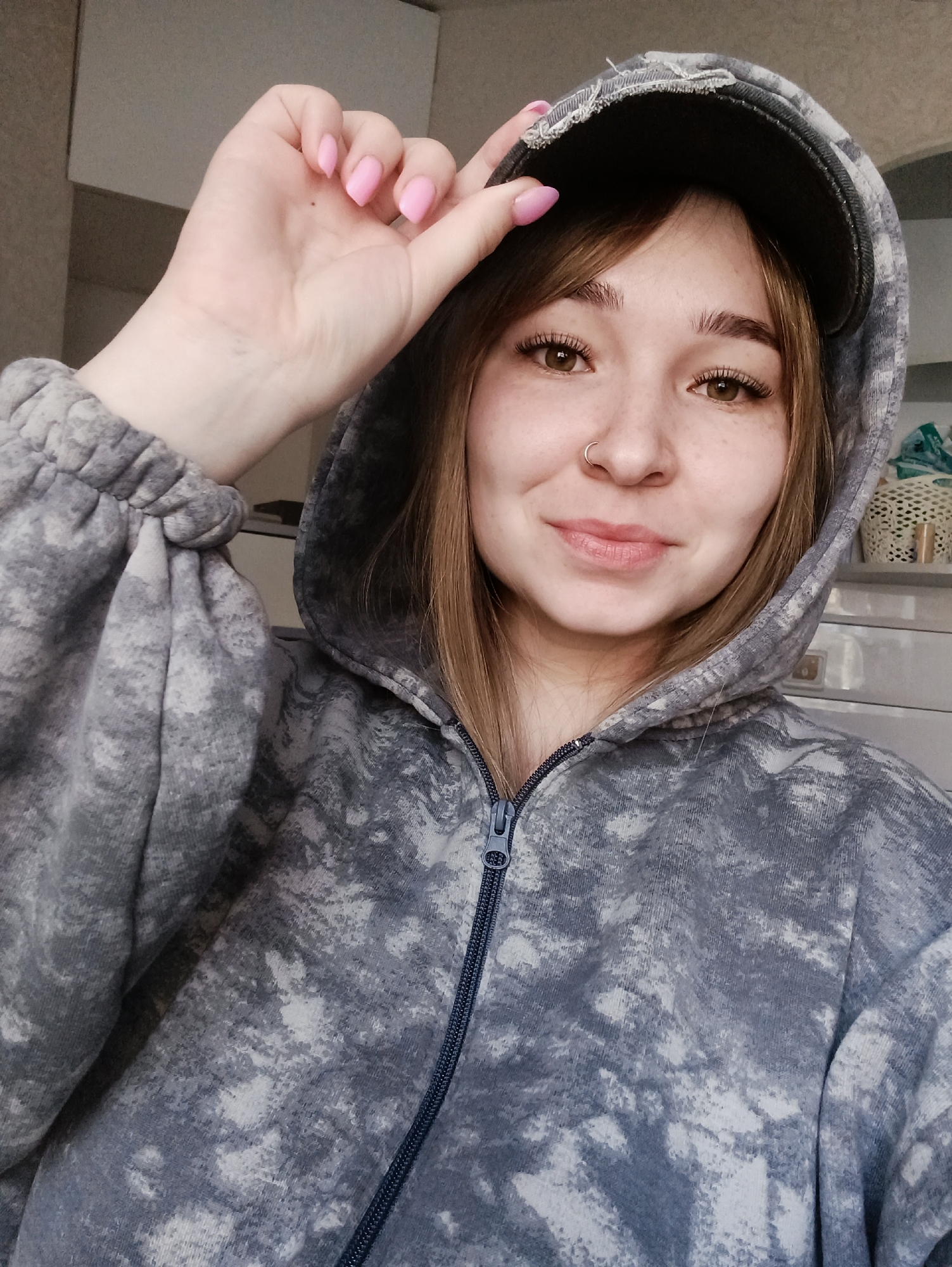 A young female subject is depicted from the chest up in an indoor setting. She has light-toned skin, light brown hair with bangs, visible freckles, and brown eyes with long eyelashes. A small, silver-colored hoop nose ring is present in her left nostril. Her lips appear natural with a slight upward curve, indicating a faint smile.
She wears a hooded sweatshirt featuring a grey and white tie-dye or bleached pattern, with the zipper partially pulled up. The hood is raised and covers her head. Beneath the hood, a dark-colored baseball cap is visible, with frayed edges on the bill. Her right hand, featuring short, pink-polished fingernails, is positioned to lightly touch the brim of the cap. The sweatshirt has elasticized cuffs, one of which is visible on her right arm.
The background consists of a plain, light-colored wall or surface, suggesting an indoor room. To the right of the subject, part of a white woven basket and a light-colored furniture piece with indistinct items are discernible. The lighting is even, illuminating the subject's face directly. The precise location within Izhevsk, Russia, is not identifiable from the visual evidence provided; the scene is consistent with a residential interior.