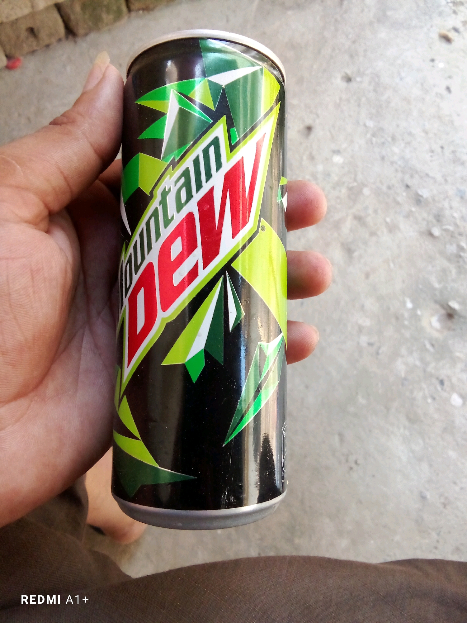 A close-up shot shows a hand holding a can of Mountain Dew. The can is black with a distinctive green and red geometric design. The background is a rough, concrete surface with some scattered pebbles, suggesting an outdoor or industrial setting. The lower portion of the image shows a section of dark brown fabric, possibly pants or a blanket. The lighting is natural, casting a slight shadow on the can. The image is geotagged to Toorabad, Pakistan.