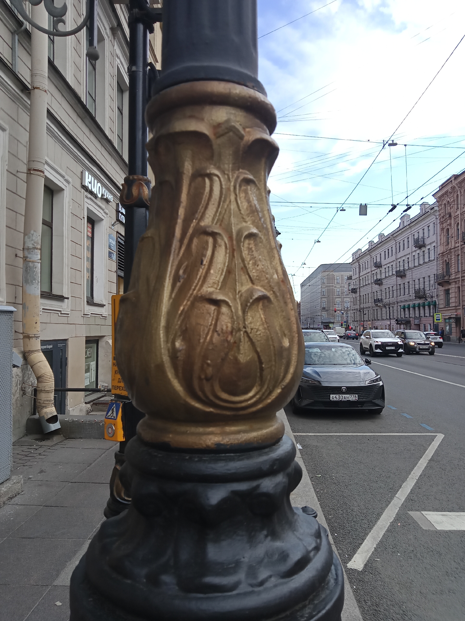 The image shows a close-up of an ornate street lamp in the foreground, with a street scene in the background. The street lamp has a dark, textured base and a golden, decorative section with intricate carvings resembling leaves or petals. In the background, a paved sidewalk is visible to the left, leading to buildings with light-colored facades and numerous windows. A white pipe runs down the side of one of the buildings. The street itself is lined with cars and buildings on the right side. The sky is overcast with a network of overhead wires. The overall impression is of an urban environment, possibly in Europe, given the architectural style and the presence of trams or trolleybuses indicated by the wires.
