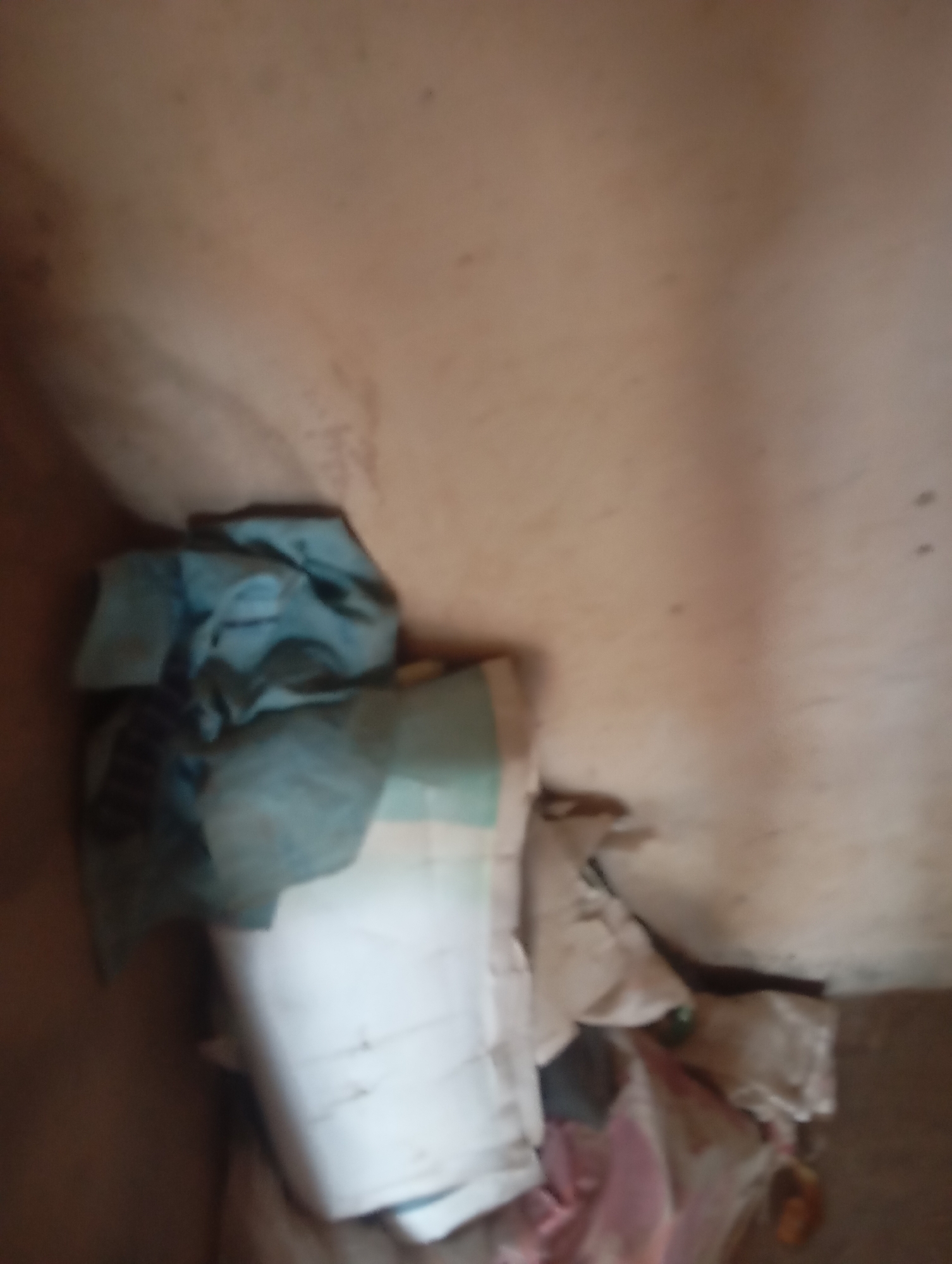 The image depicts a pile of laundry, predominantly white and blue fabrics, resting against a pale, textured wall. The lighting is dim, casting soft shadows and obscuring finer details. The scene appears to be indoors, possibly a domestic setting within Jauro Yinu, Nigeria, suggested by the cultural context. No individuals are visible in the image. The laundry is crumpled and unevenly stacked, with some items appearing to be folded or loosely bundled. The wall behind the laundry is slightly grimy and shows signs of age.